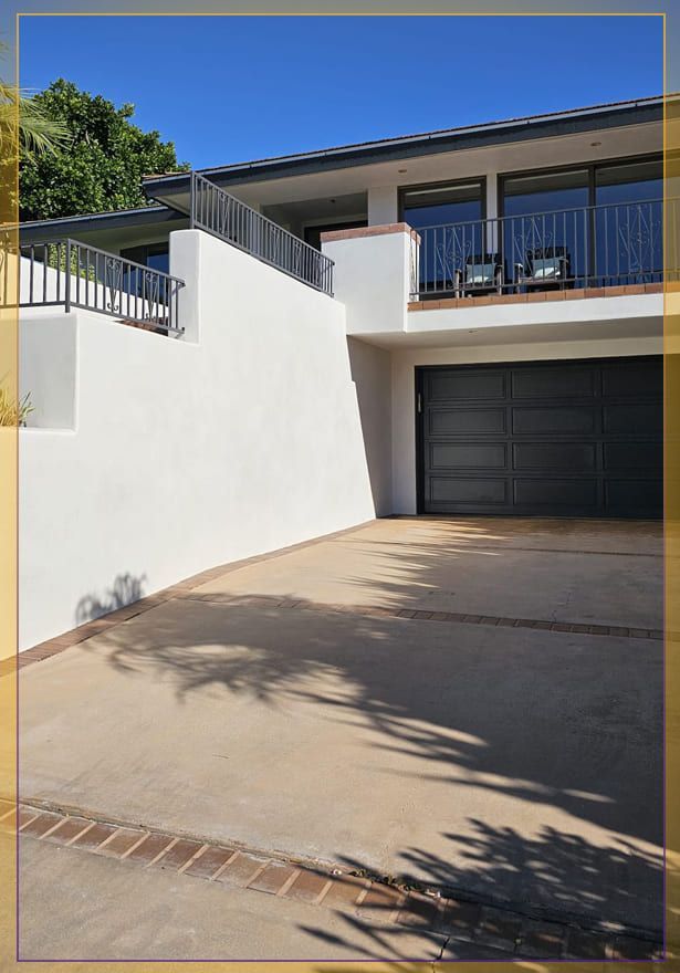 Driveway leading to a white house with a dark garage door and balcony under a blue sky