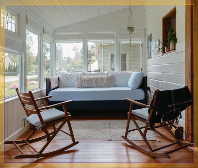 Bright sunroom with a gray daybed, wooden chairs, and a black fireplace on a hardwood floor.