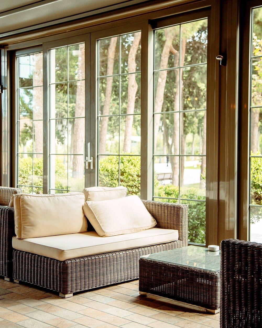 Sunlit patio with wicker sofa, ottoman, and large windows overlooking greenery