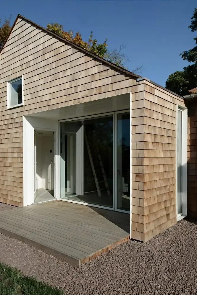 Modern small house with wood shingles, large glass doors, and a gravel patio under a sloped roof