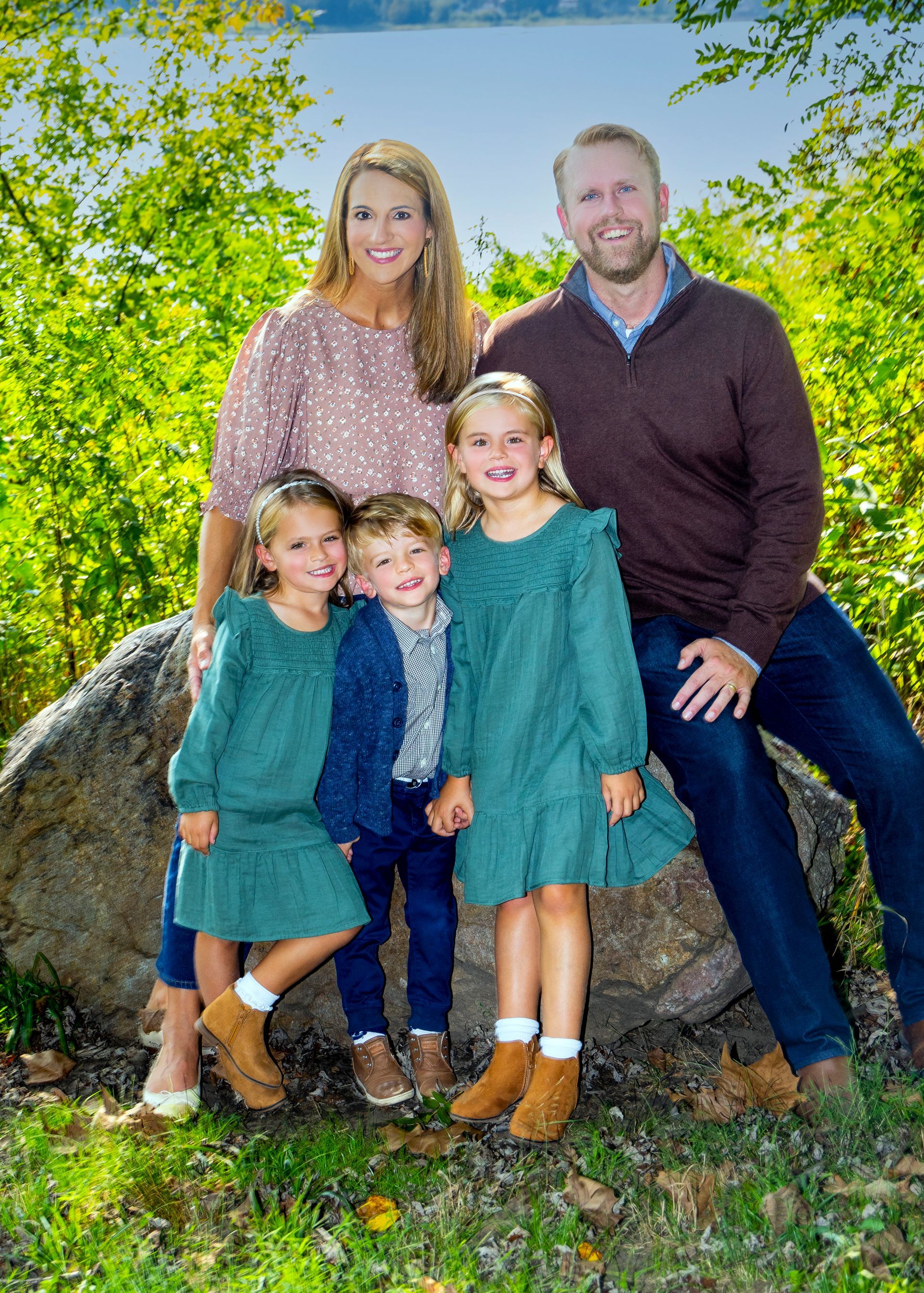 Family of five posing outdoors in greenery, smiling and dressed in teal and brown outfits.