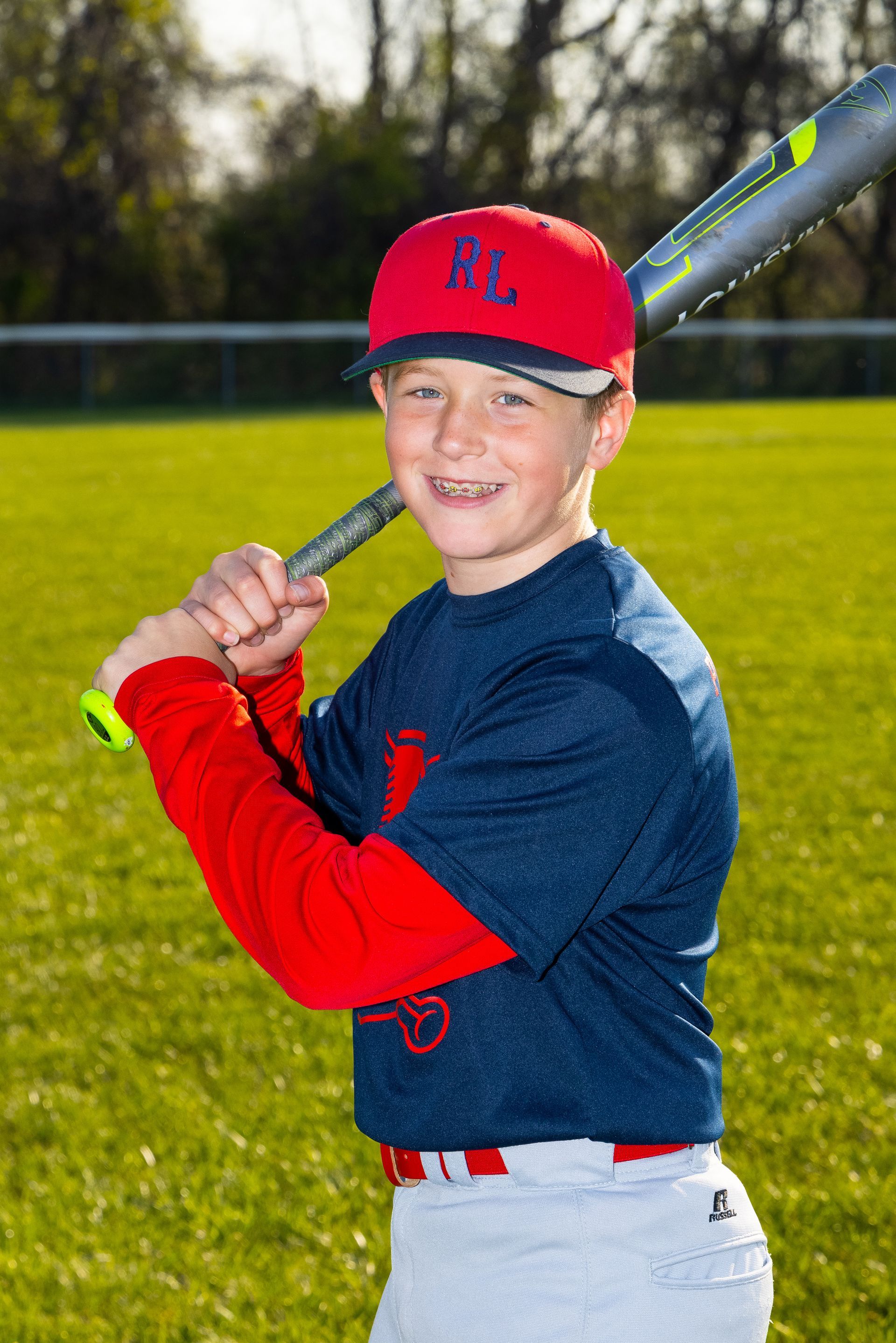Boy in red cap holding a baseball bat on a field, wearing a navy shirt and red sleeves, smiling at the camera