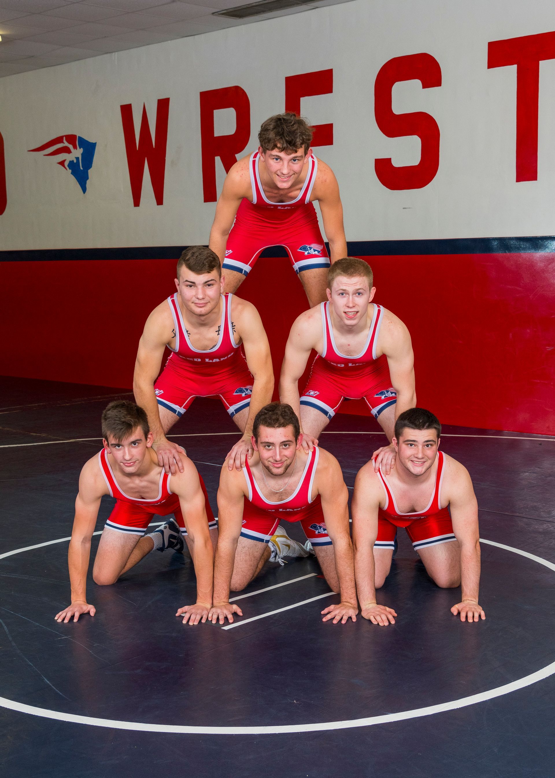 Six wrestlers in red singlets posing on a wrestling mat in a gymnasium