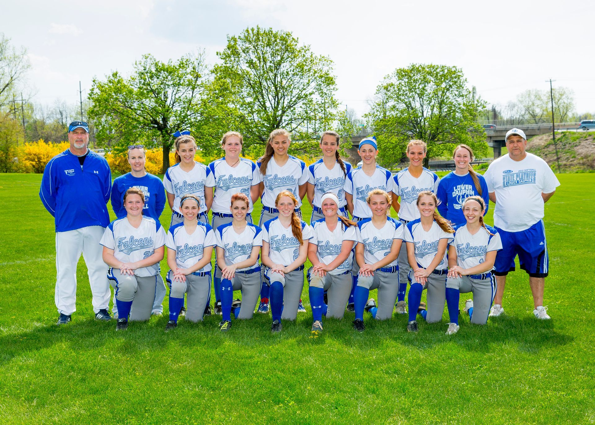 Baseball team posing on a grassy field, wearing white and blue uniforms with two coaches on the ends