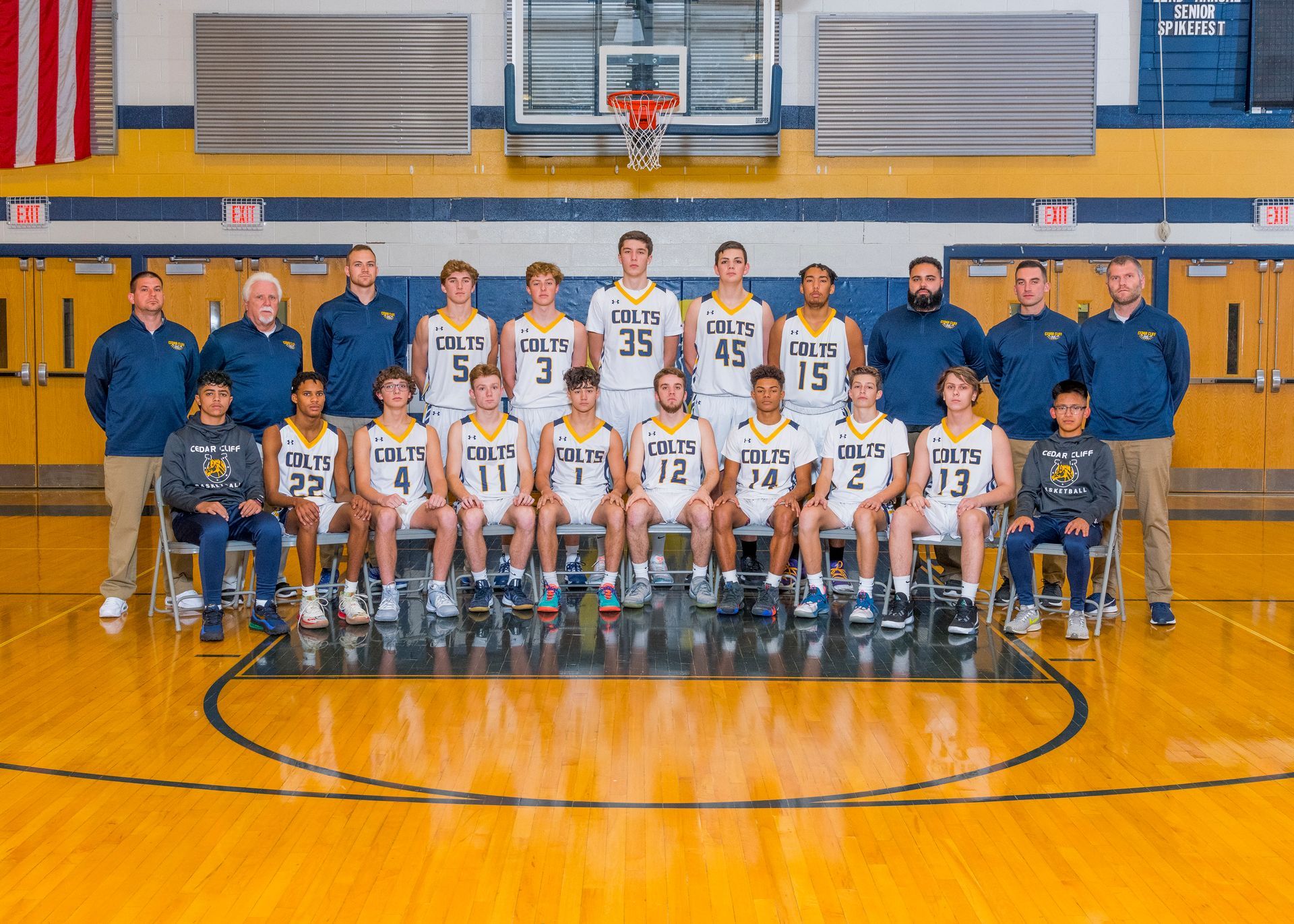 Group of basketball players and coaches posing for a team photo in a gymnasium with orange floor and blue walls.