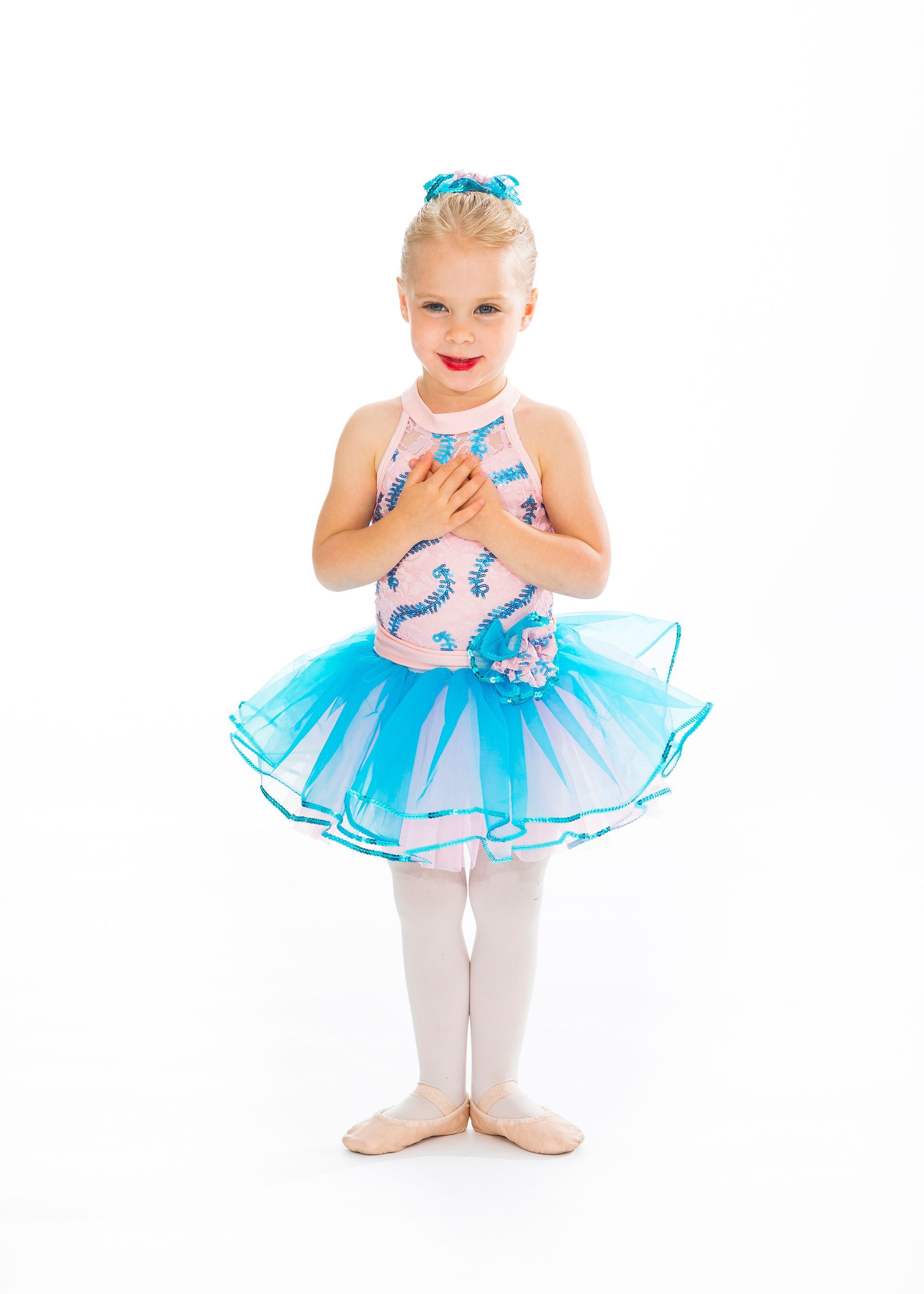 Young child in a blue tutu and bows, standing against a white background, smiling with hands clasped.