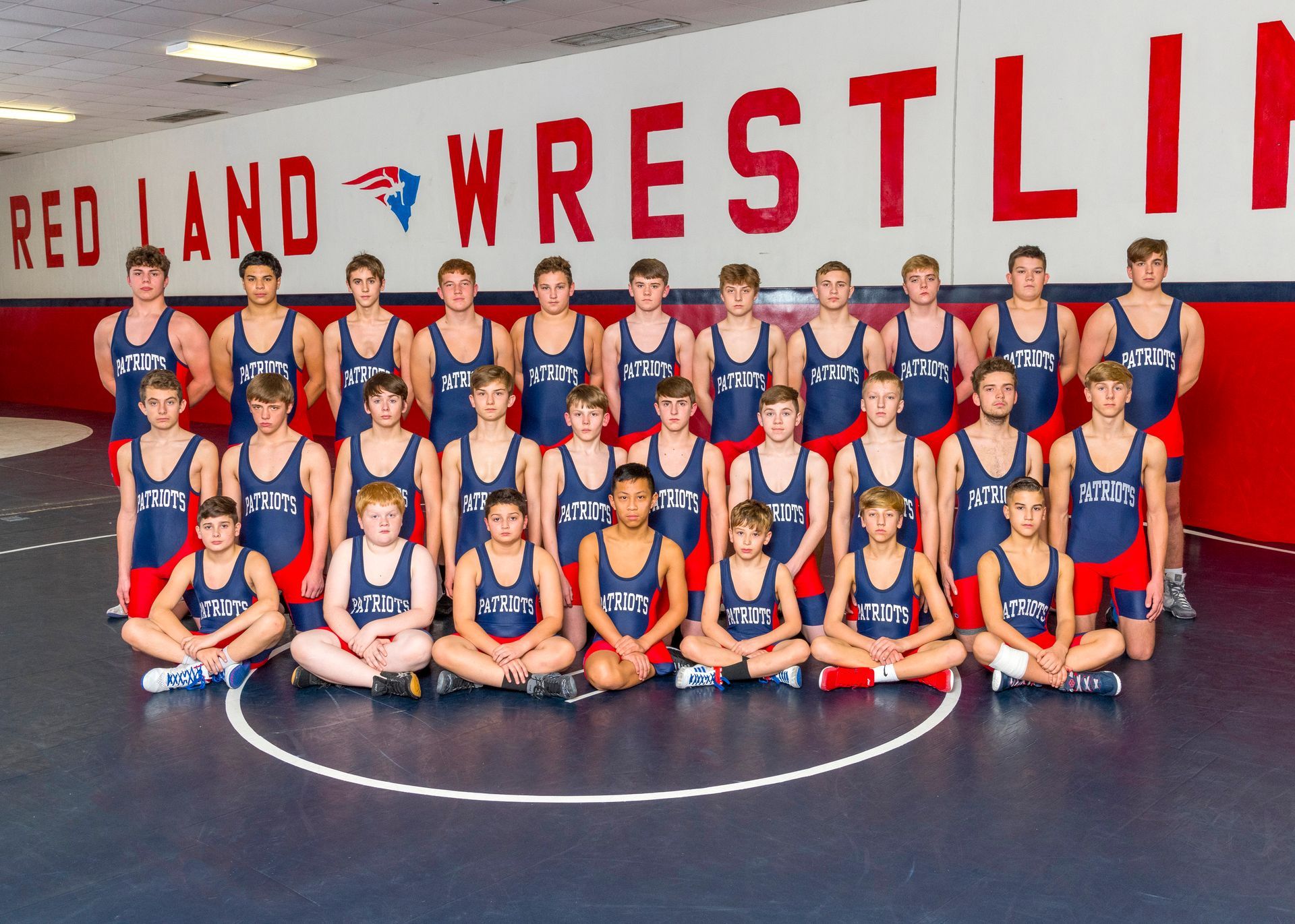 Wrestling team group photo in red and blue singlets posing on a mat under a “Red Land Wrestling” banner