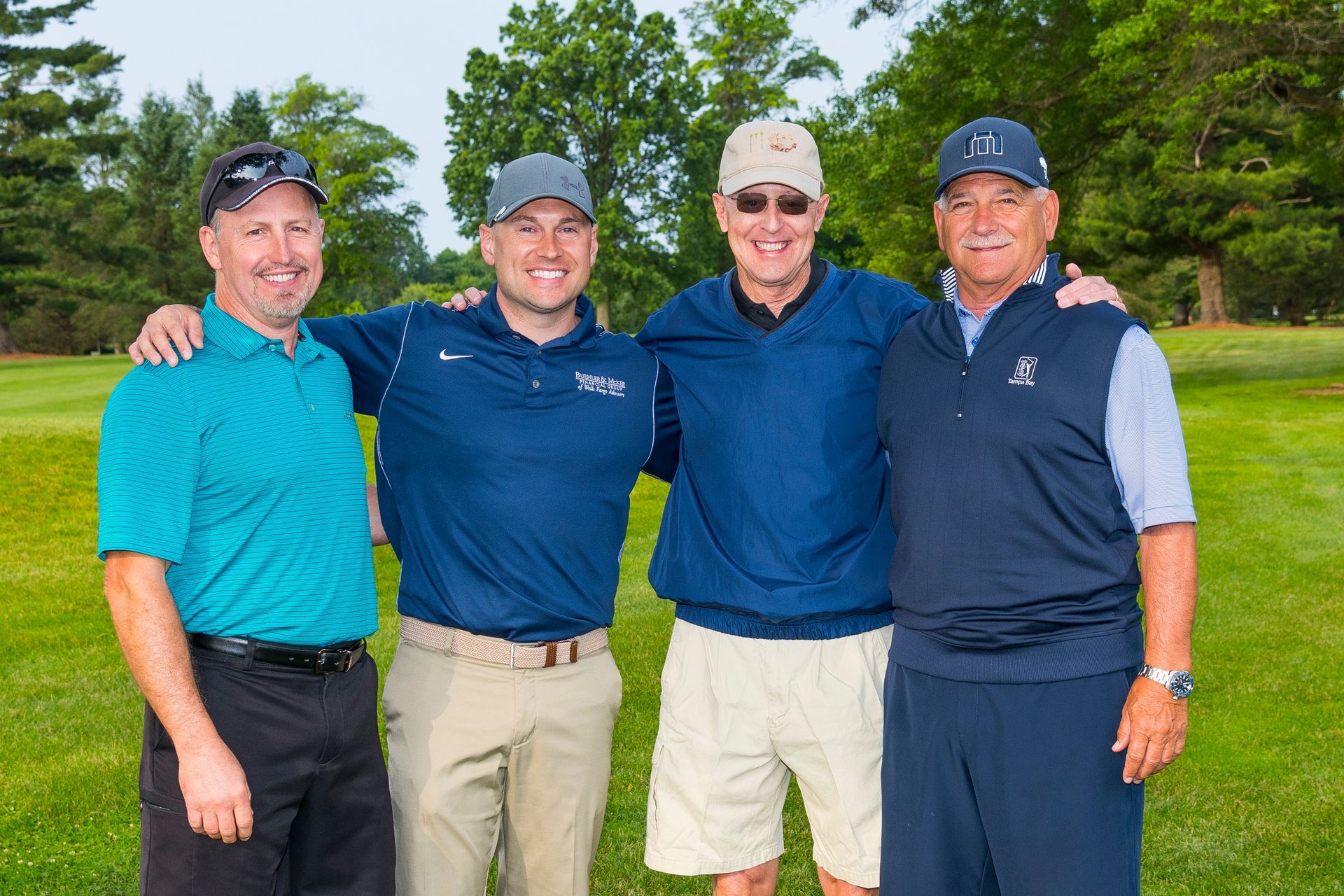 Four friends posing outdoors on a golf course, smiling with arms around each other.