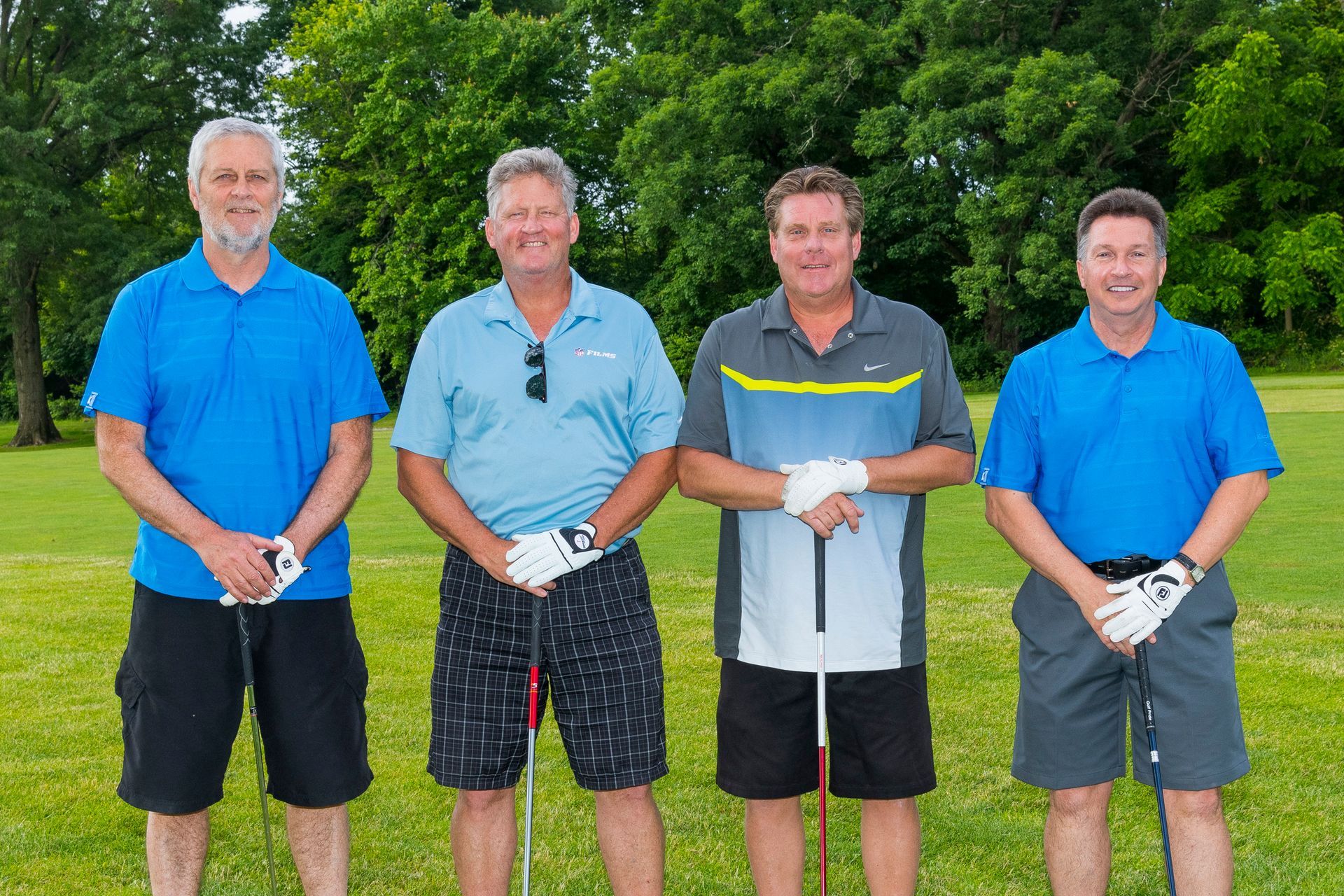Four men stand outdoors on grass, holding golf clubs with trees in the background.
