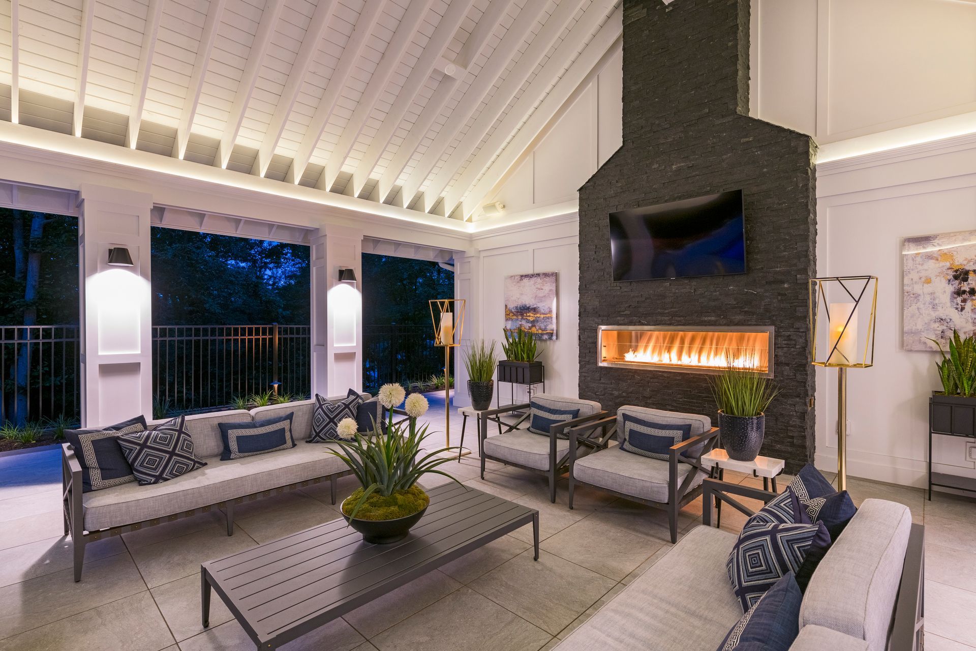 Modern living room with fireplace, gray sofas, patterned chairs, and tall windows at dusk