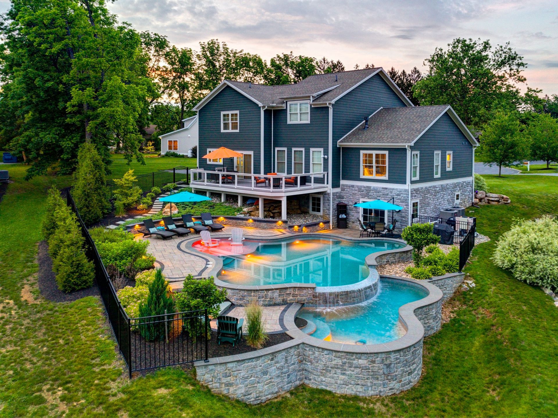 A large gray house with a backyard pool, patio umbrellas, and stone retaining walls at sunset