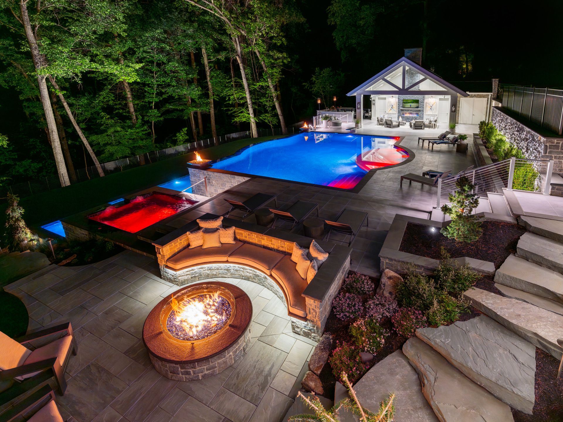 Backyard patio with illuminated pool, fire pit, lounge seating, and a lit house at night