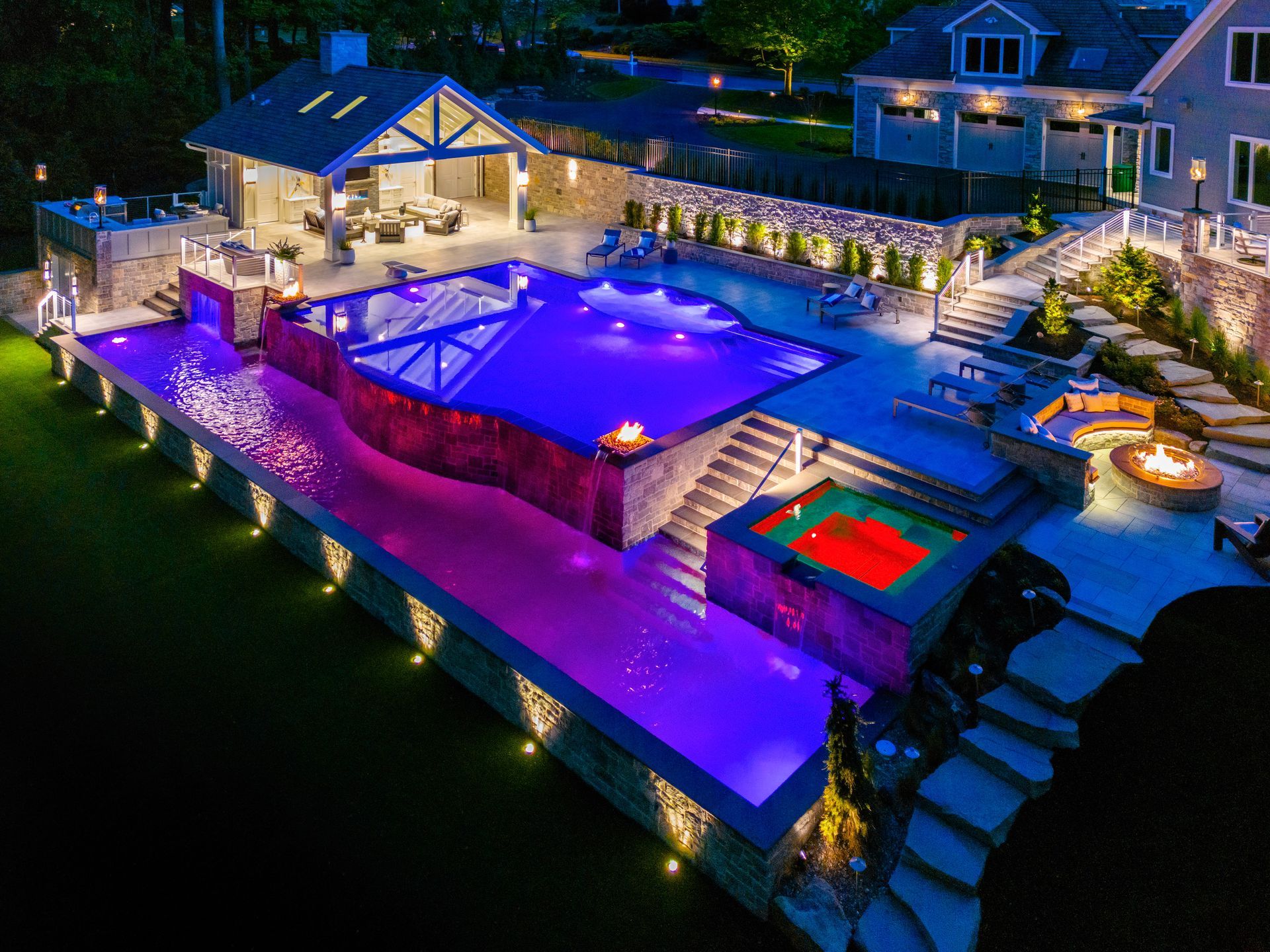 Luxury backyard with illuminated pool, purple lighting, hot tub, and patio at night