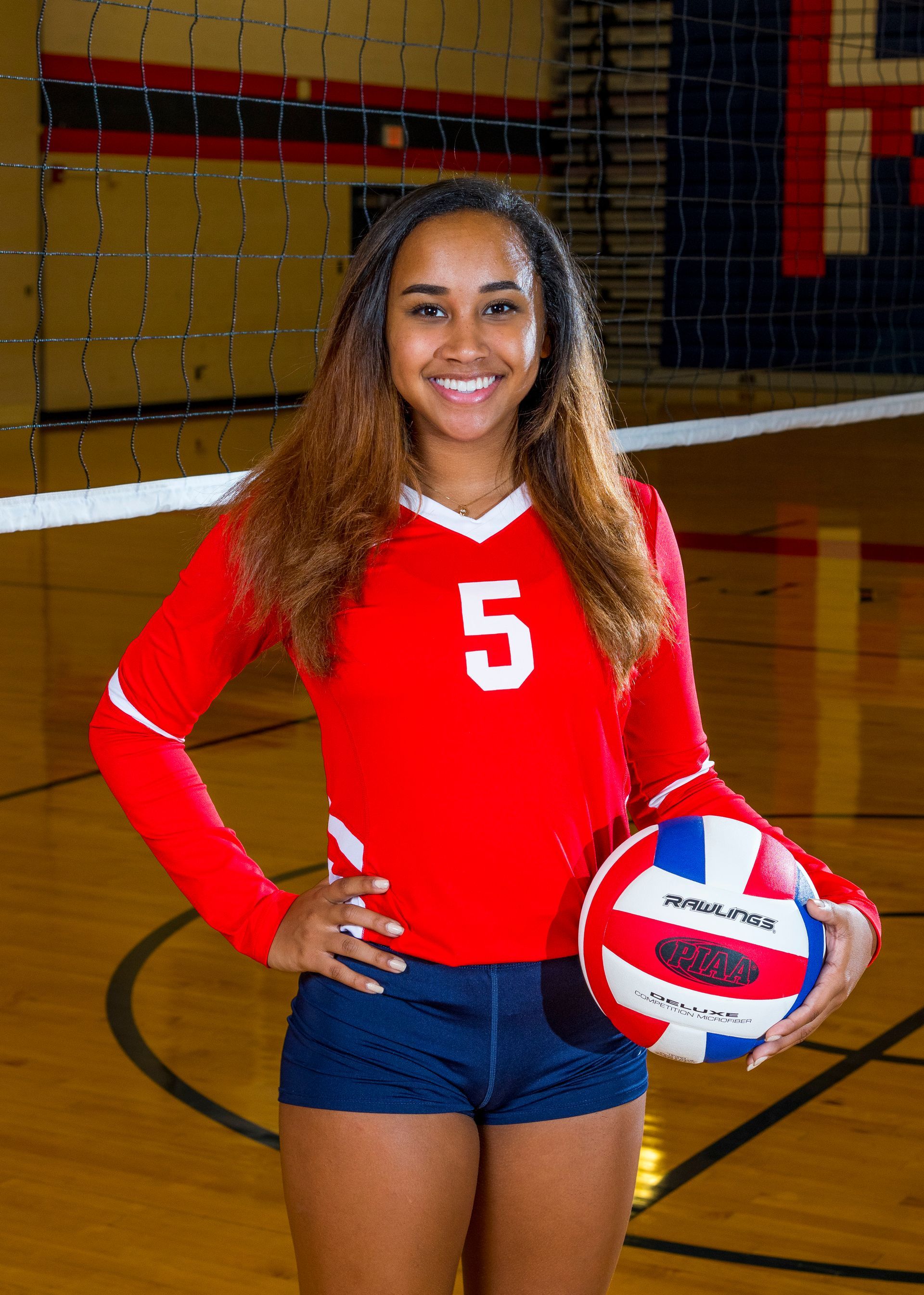 Female volleyball player in red jersey holding a volleyball on an indoor court, smiling at the camera.