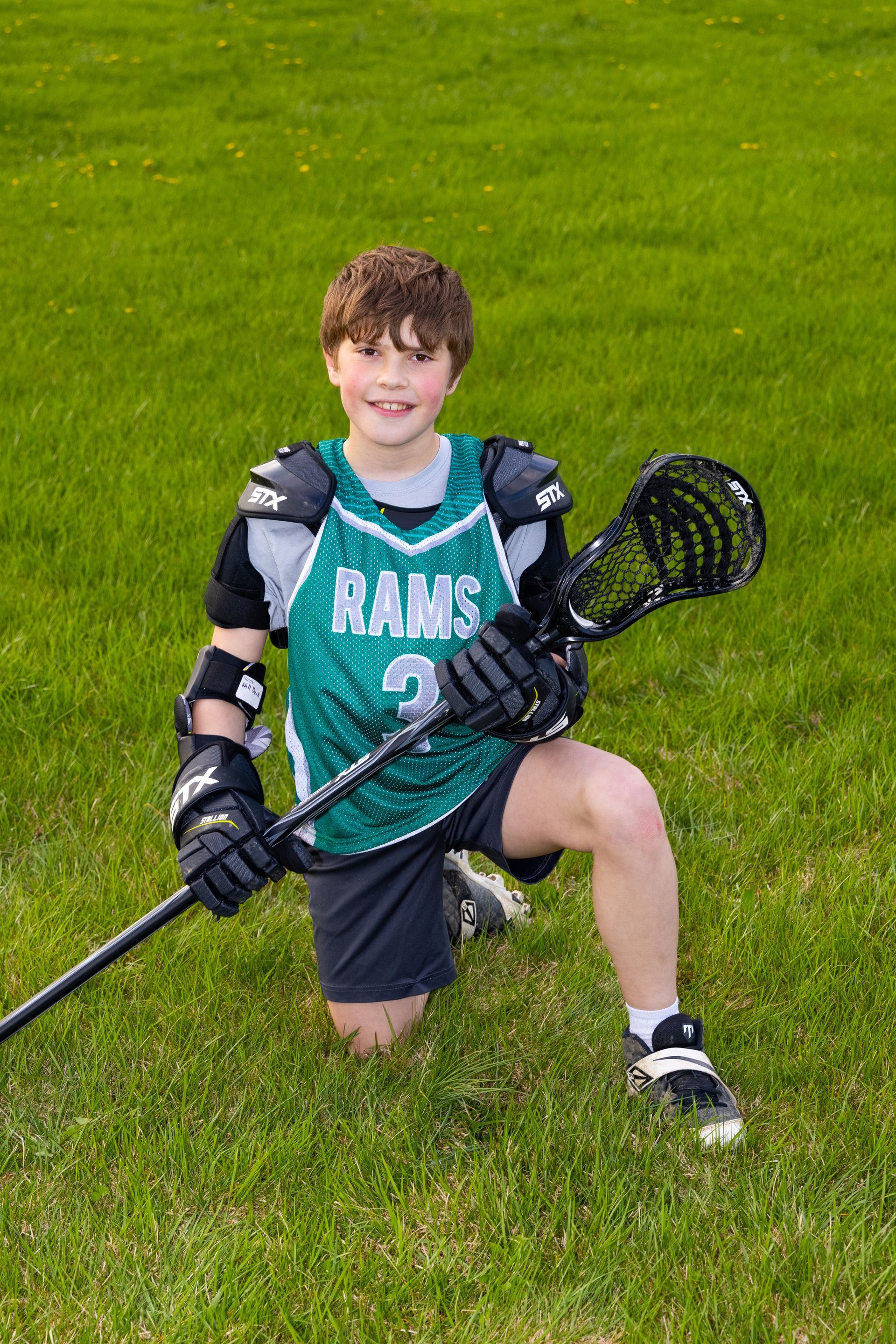Boy in teal lacrosse jersey kneeling on grass and holding a black lacrosse stick