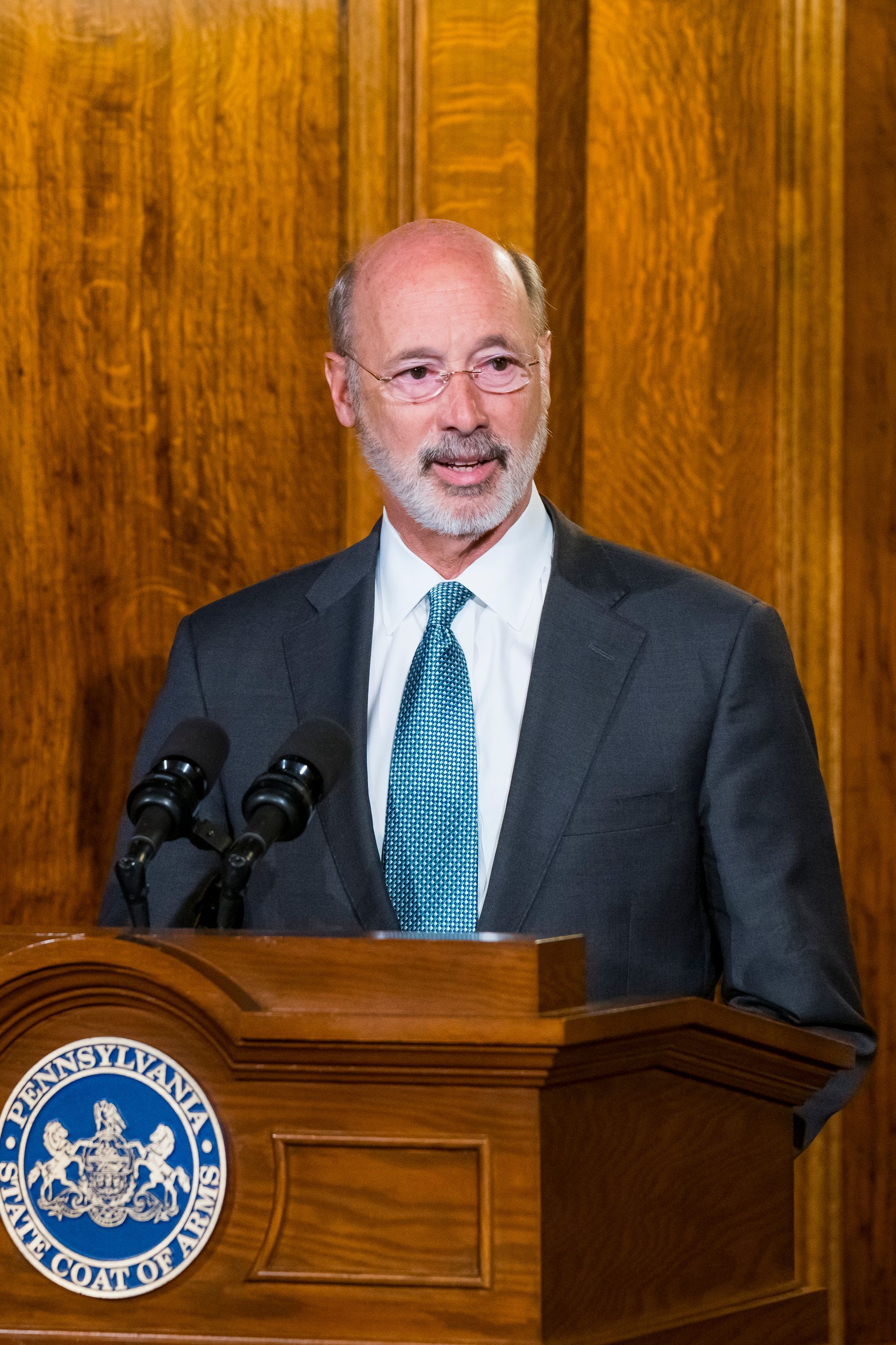 Man speaking at a podium with microphones, in a dark suit and blue tie, against a wood-paneled wall.