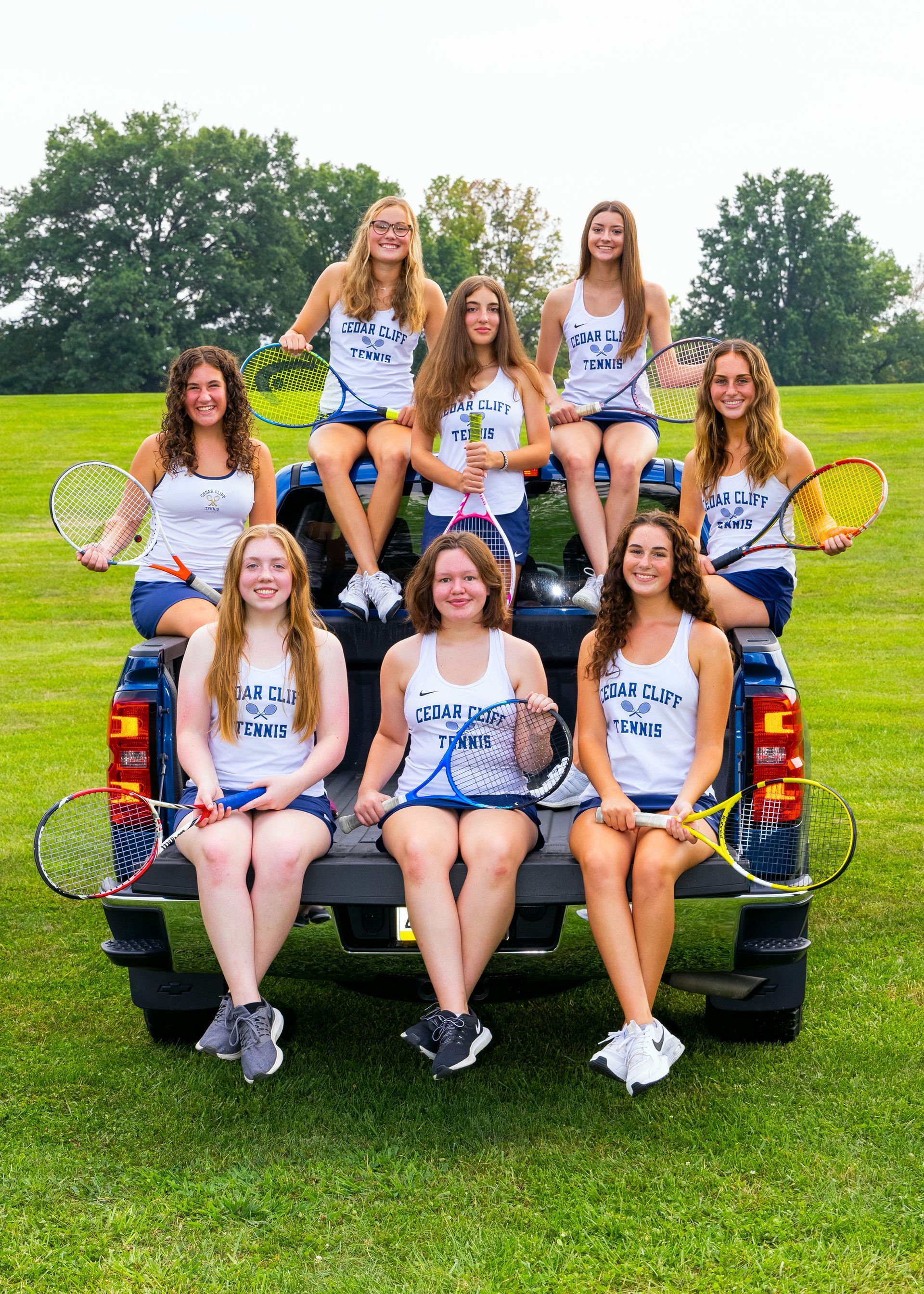 Group posed on a pickup truck in a grassy field, holding tennis rackets and wearing matching shirts.