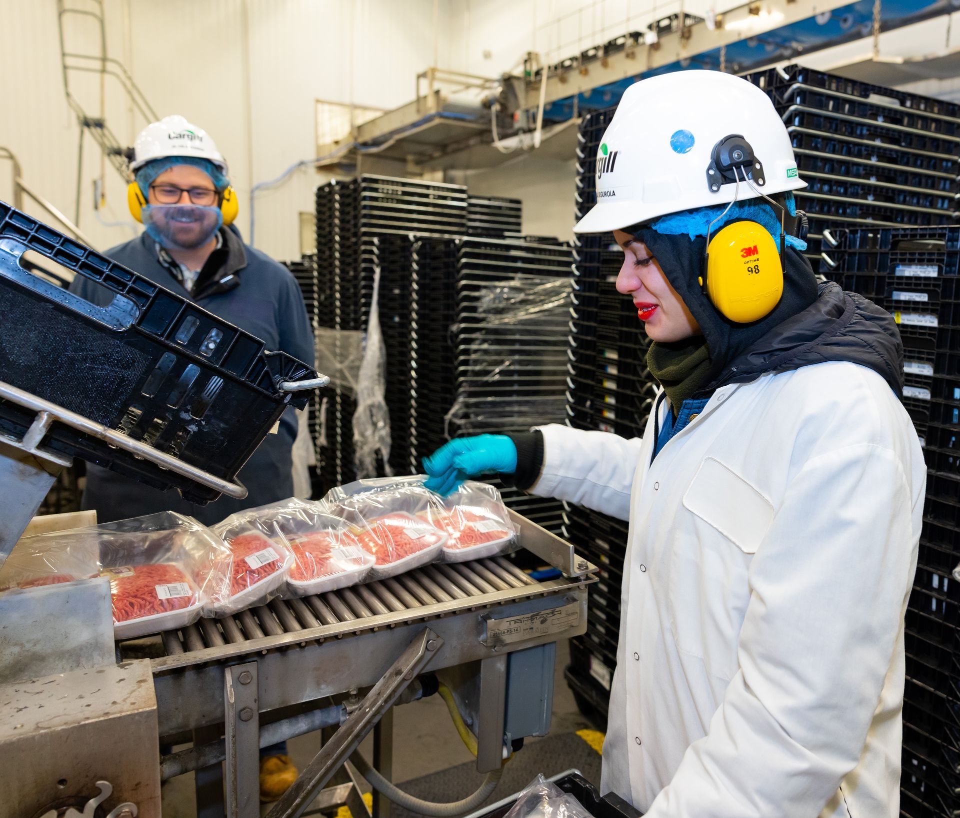 Workers packaging food in a factory, wearing safety gear beside stacks of trays.