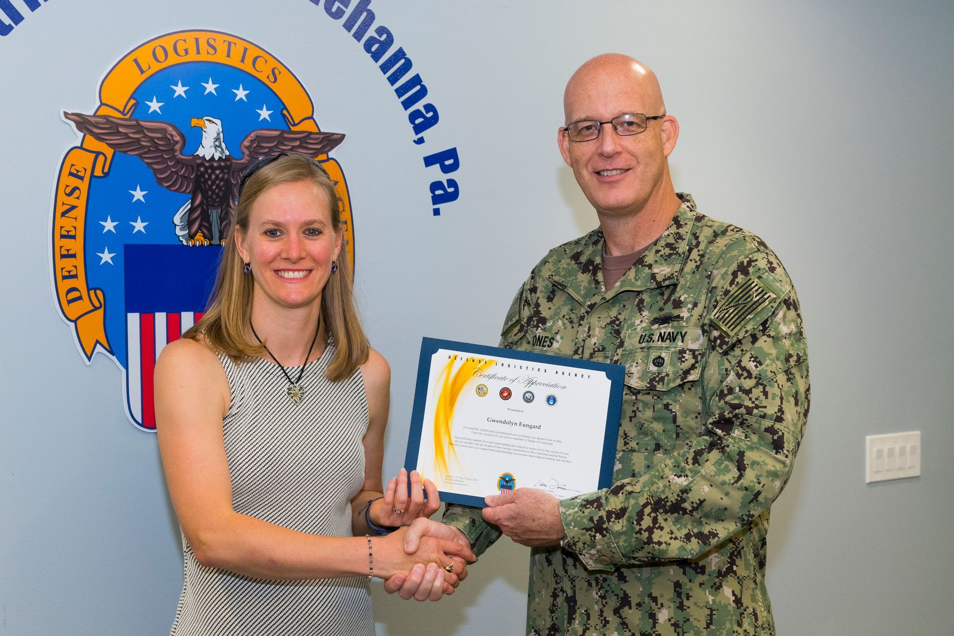 Woman receives a certificate from a uniformed man in front of a military emblem and wall logo.