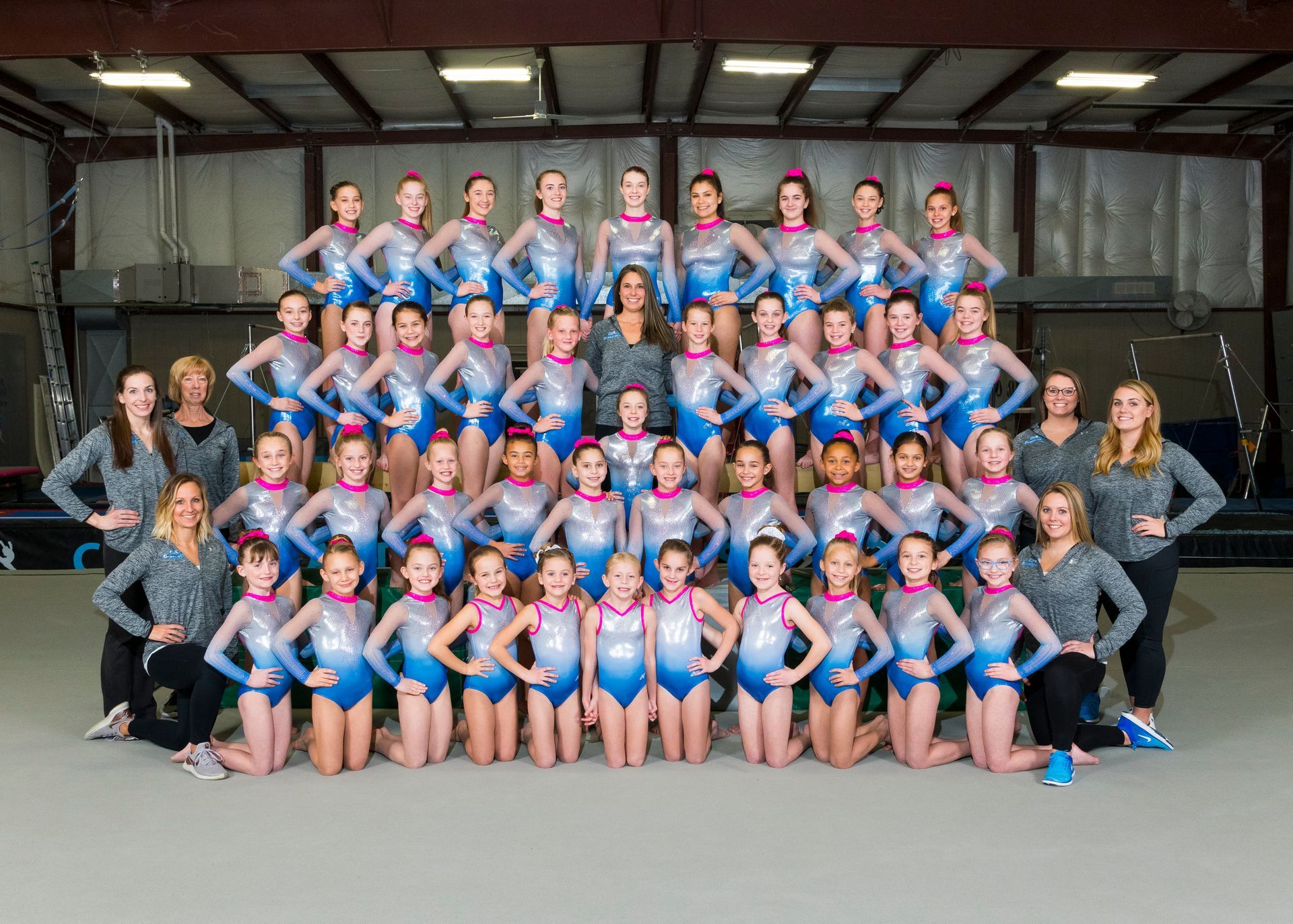 Large dance team posed in pyramid formation in a warehouse, wearing blue and pink costumes with coaches on the sides