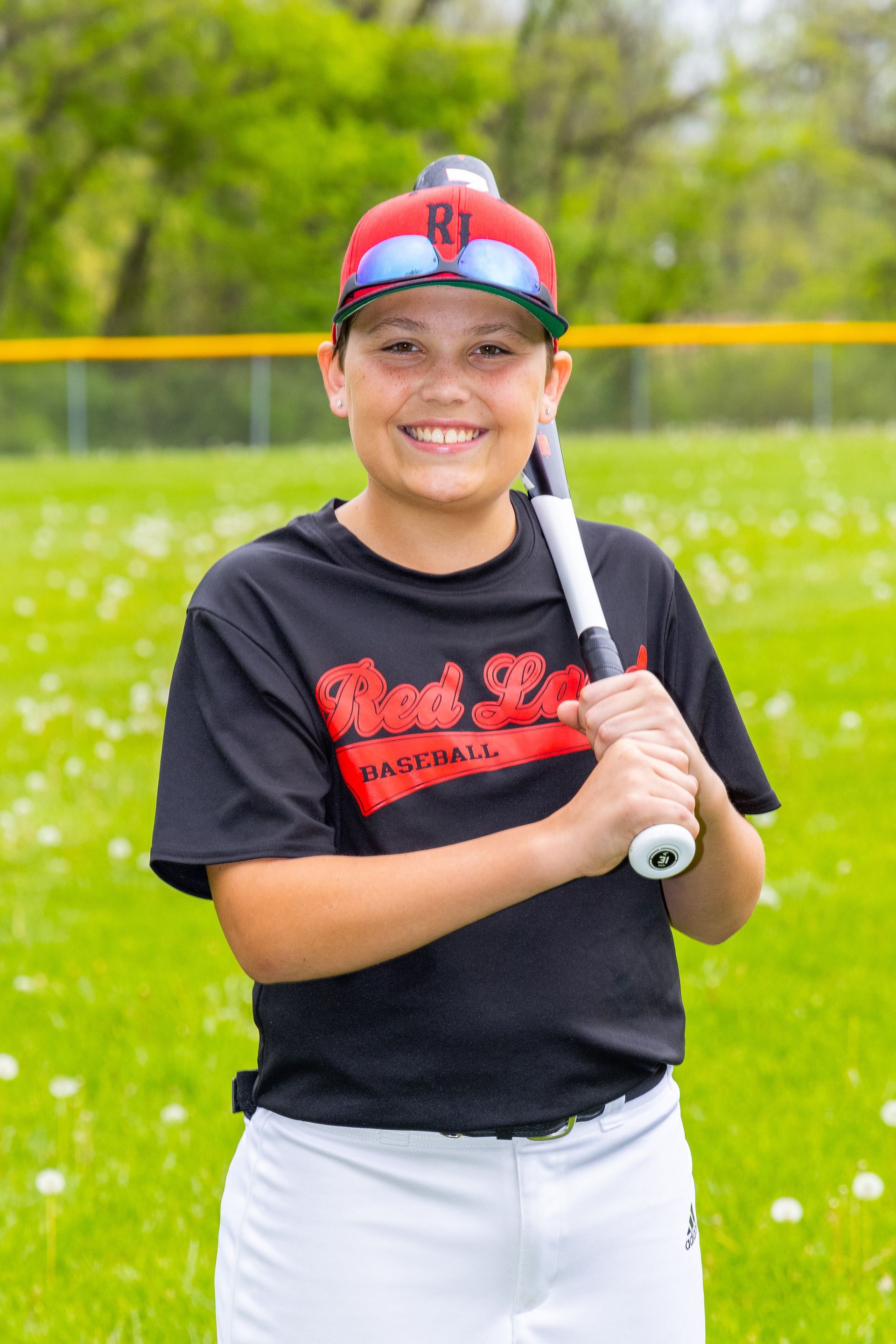 Smiling child holding a baseball bat on a grassy field, wearing a black shirt, white pants, and red cap.