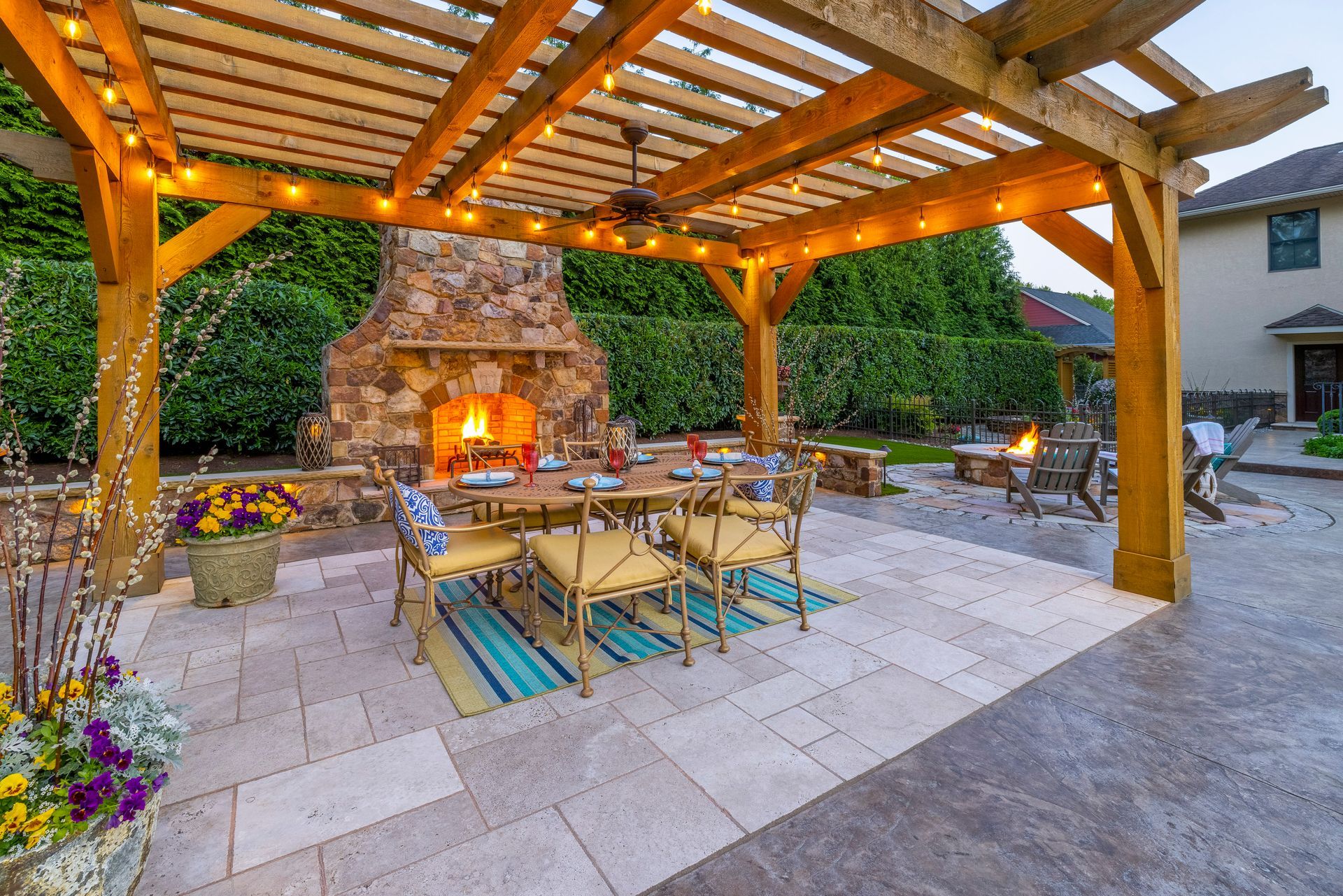 Outdoor patio with pergola, dining table, stone fireplace, and warm string lights at dusk