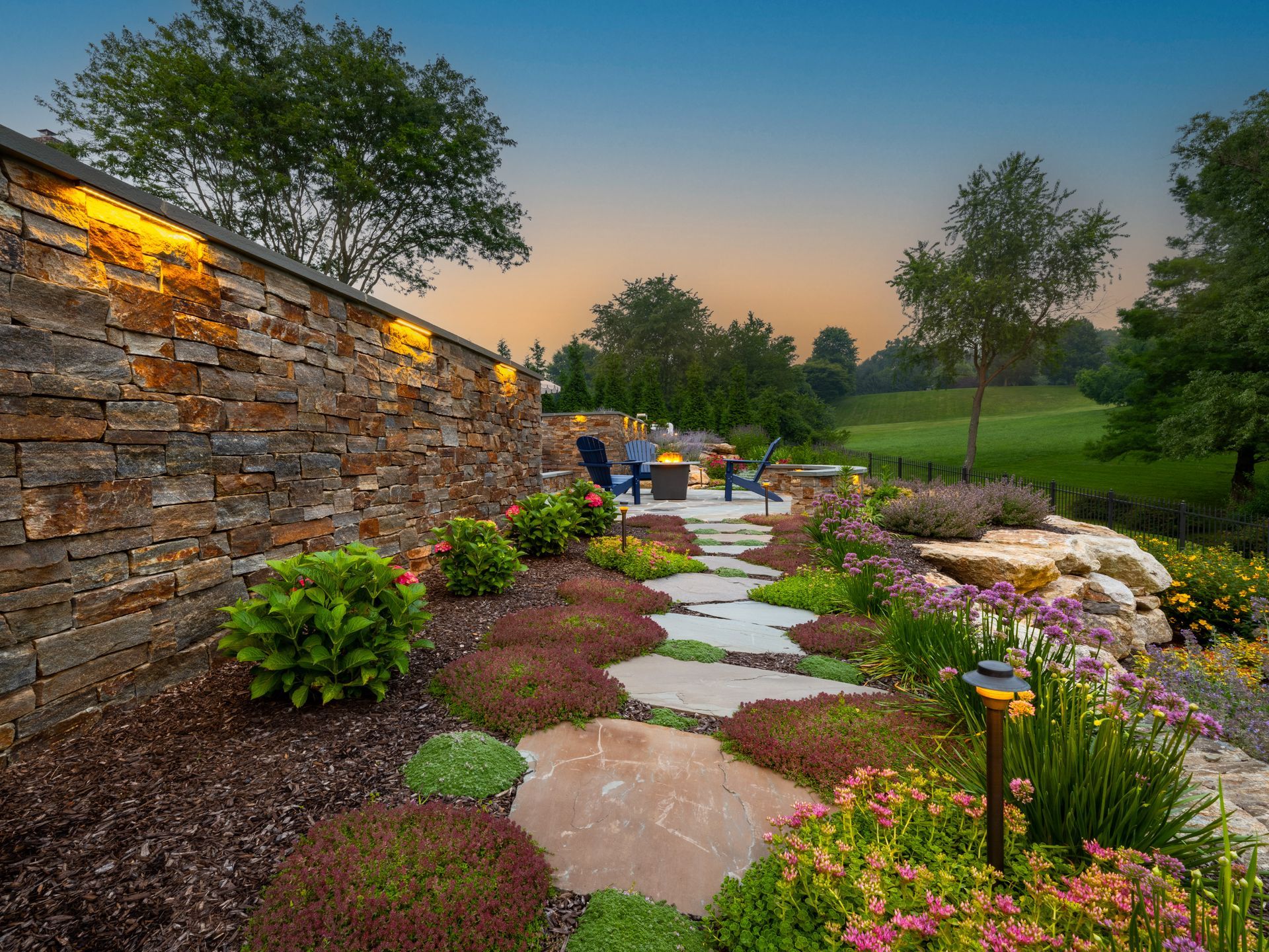 Stone garden path beside a lit retaining wall, leading through colorful flowers at dusk