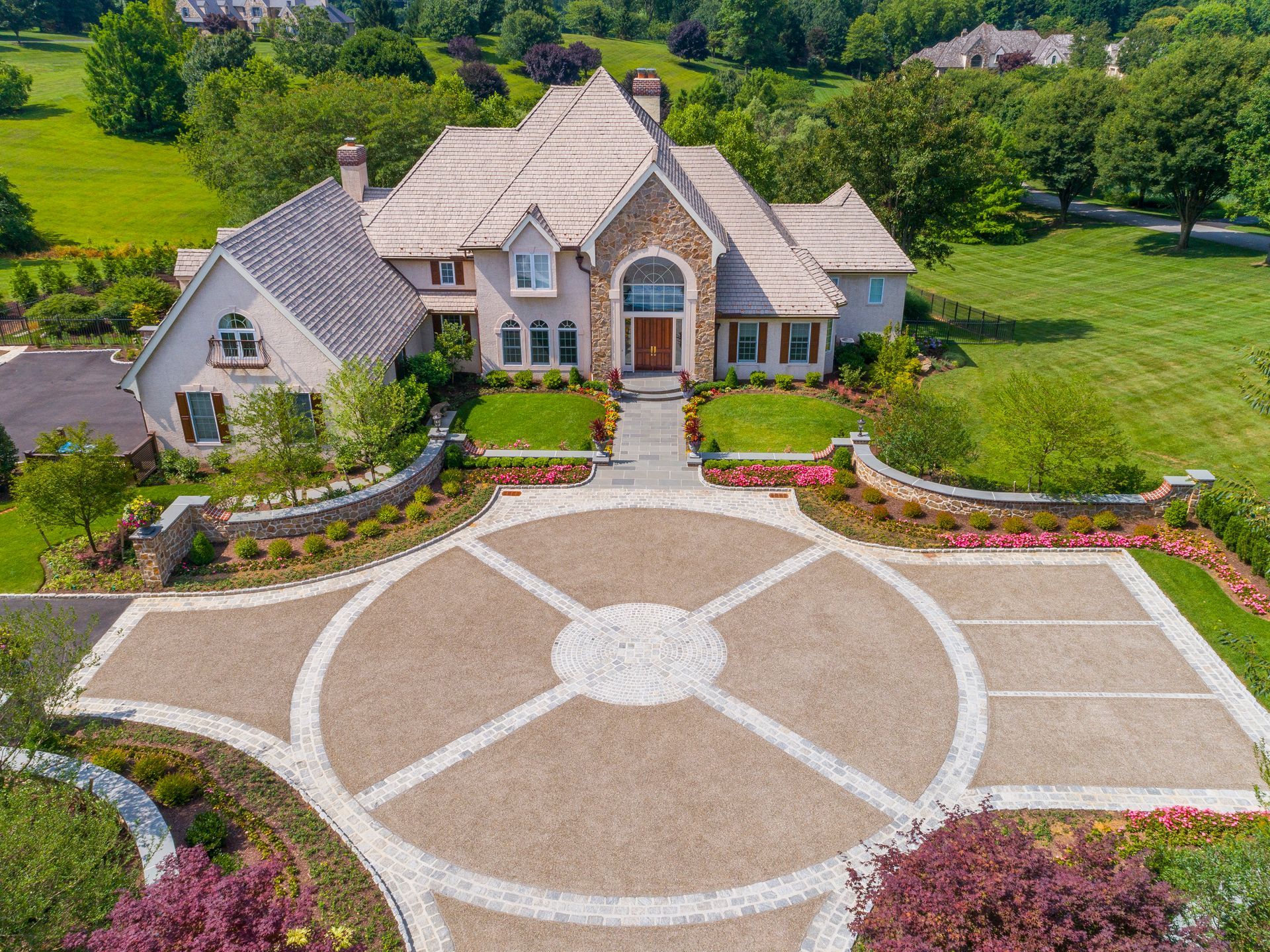 Large stone house with circular driveway, landscaped garden, and green lawn aerial view