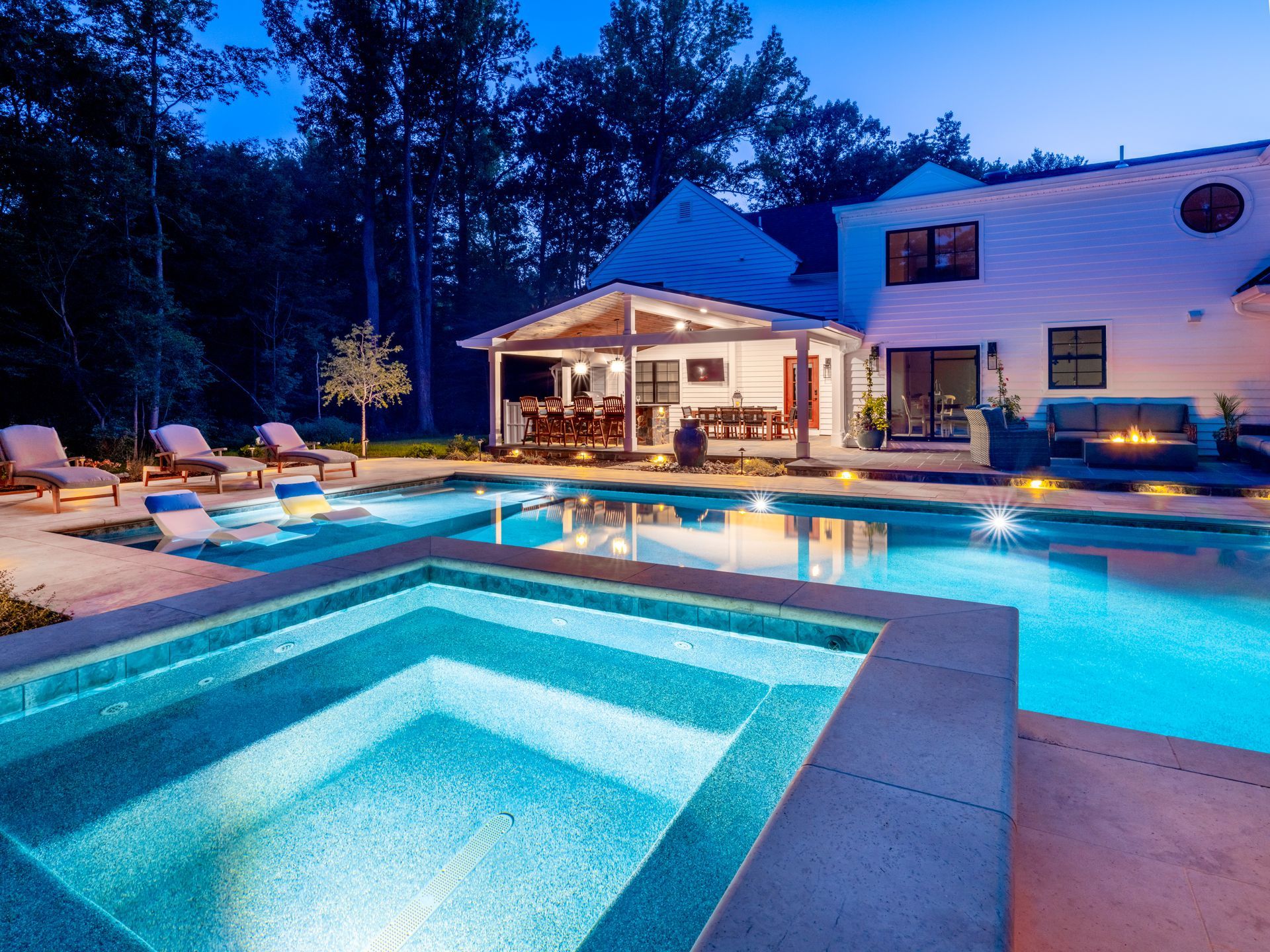 Modern backyard with lit swimming pool, hot tub, and patio beside a white house at dusk