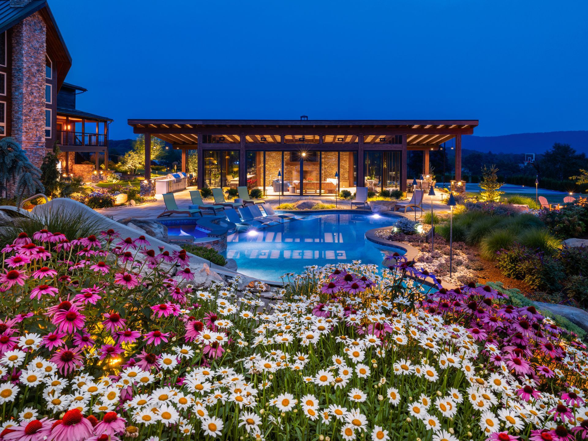 Luxury backyard with lit pool, modern patio, and colorful flower garden at dusk