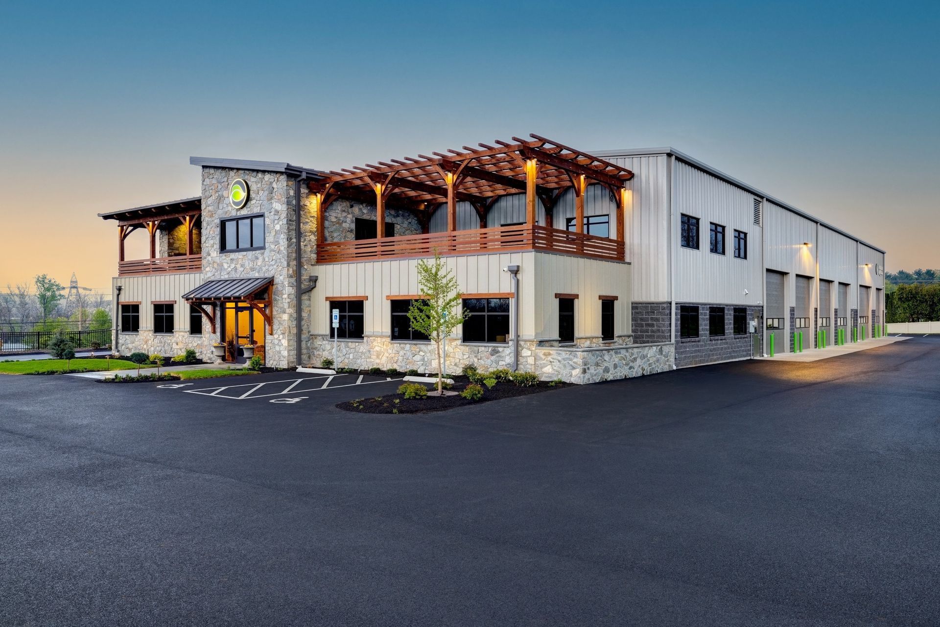 Modern two-story commercial building with stone facade, wooden pergola entrance, and large parking lot at dusk