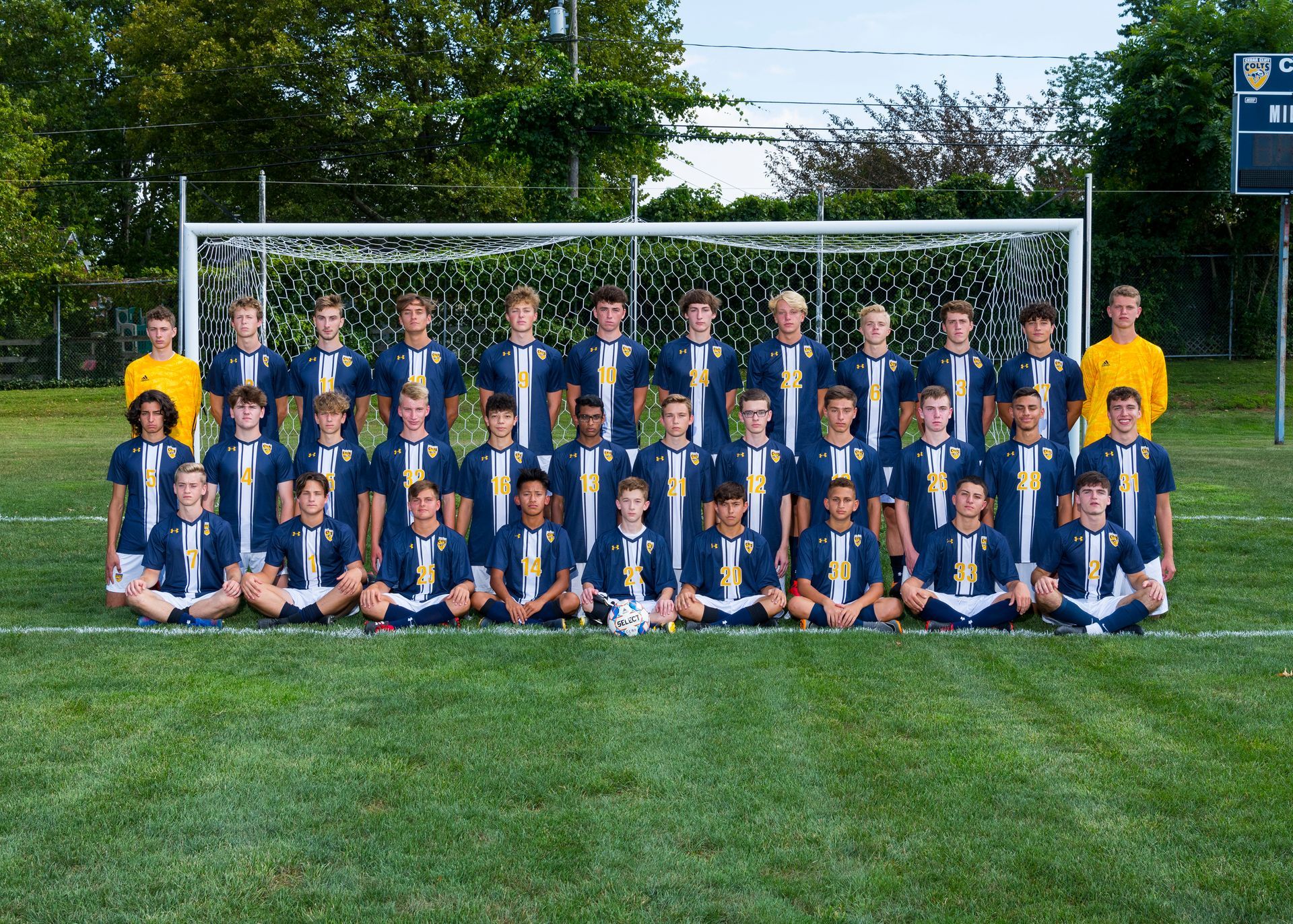 Youth soccer team posing in two rows on a field, with a goal and trees in the background.