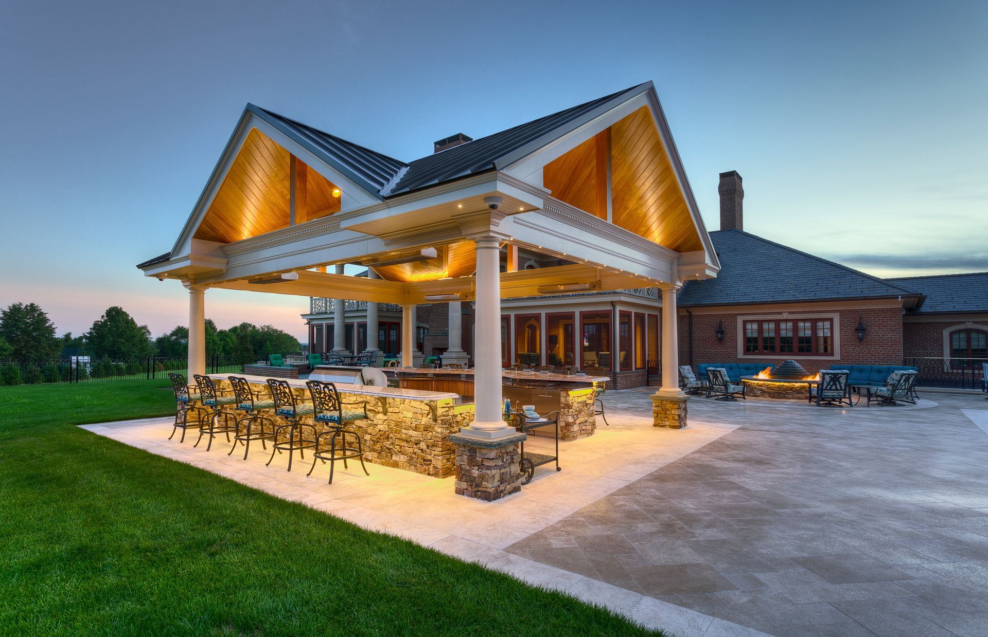 Covered outdoor patio at dusk with lit wooden beams, stone columns, and dining tables beside a modern brick home