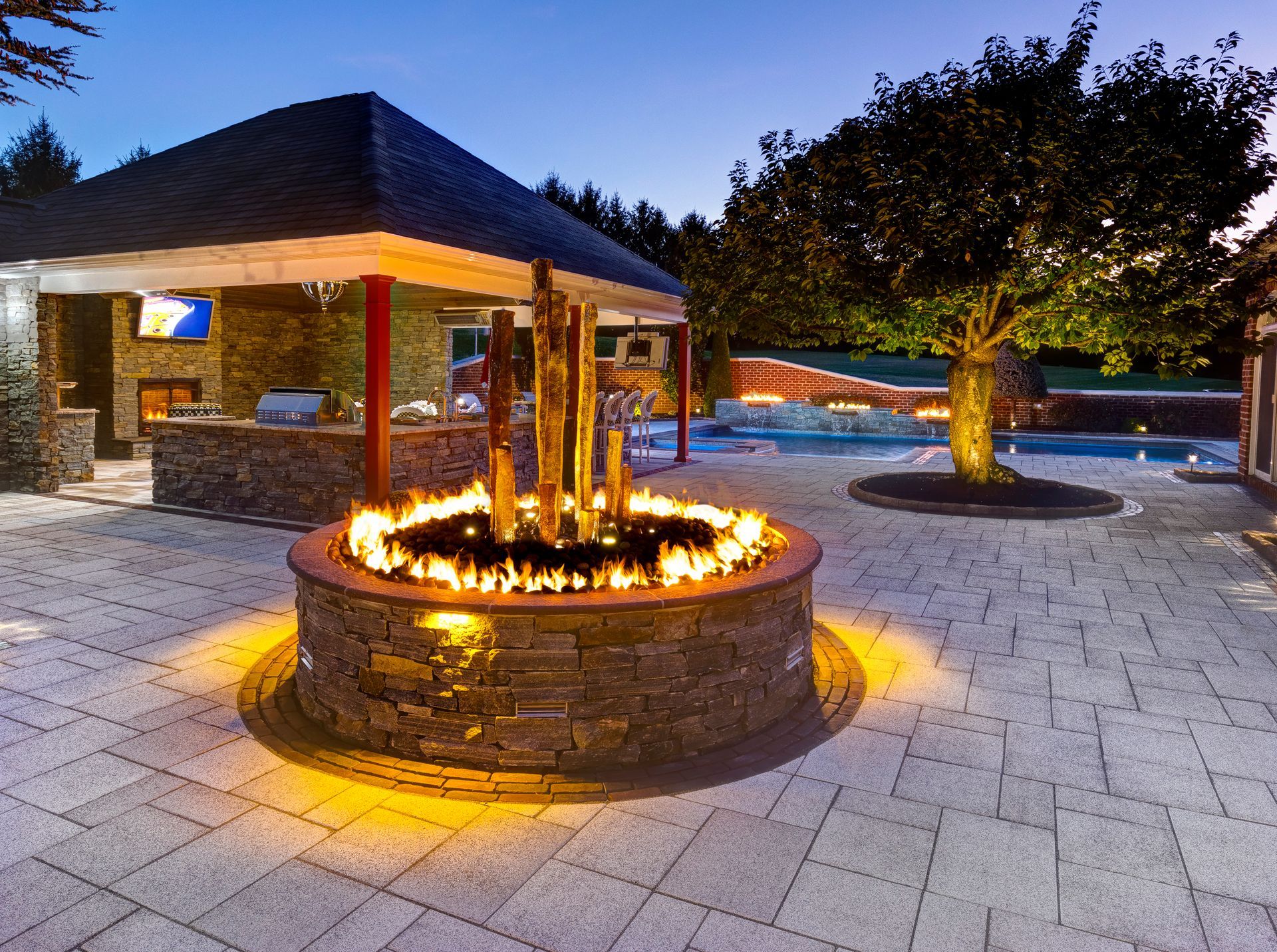 Outdoor stone fire pit glowing at dusk beside a pavilion and tree-lit paved courtyard