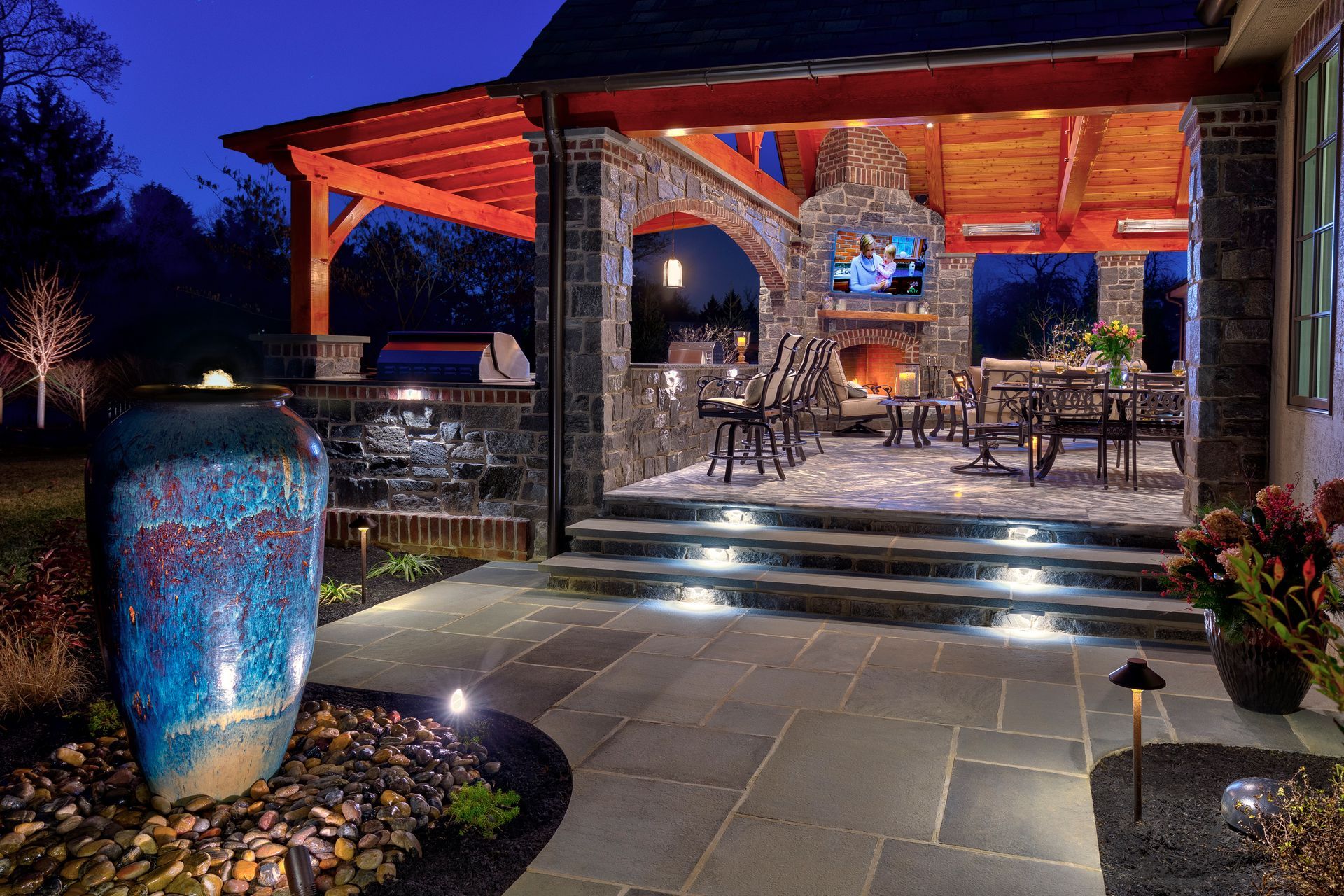 Lit stone patio with steps leading to a warmly lit porch at dusk, beside a large blue vase and flower beds