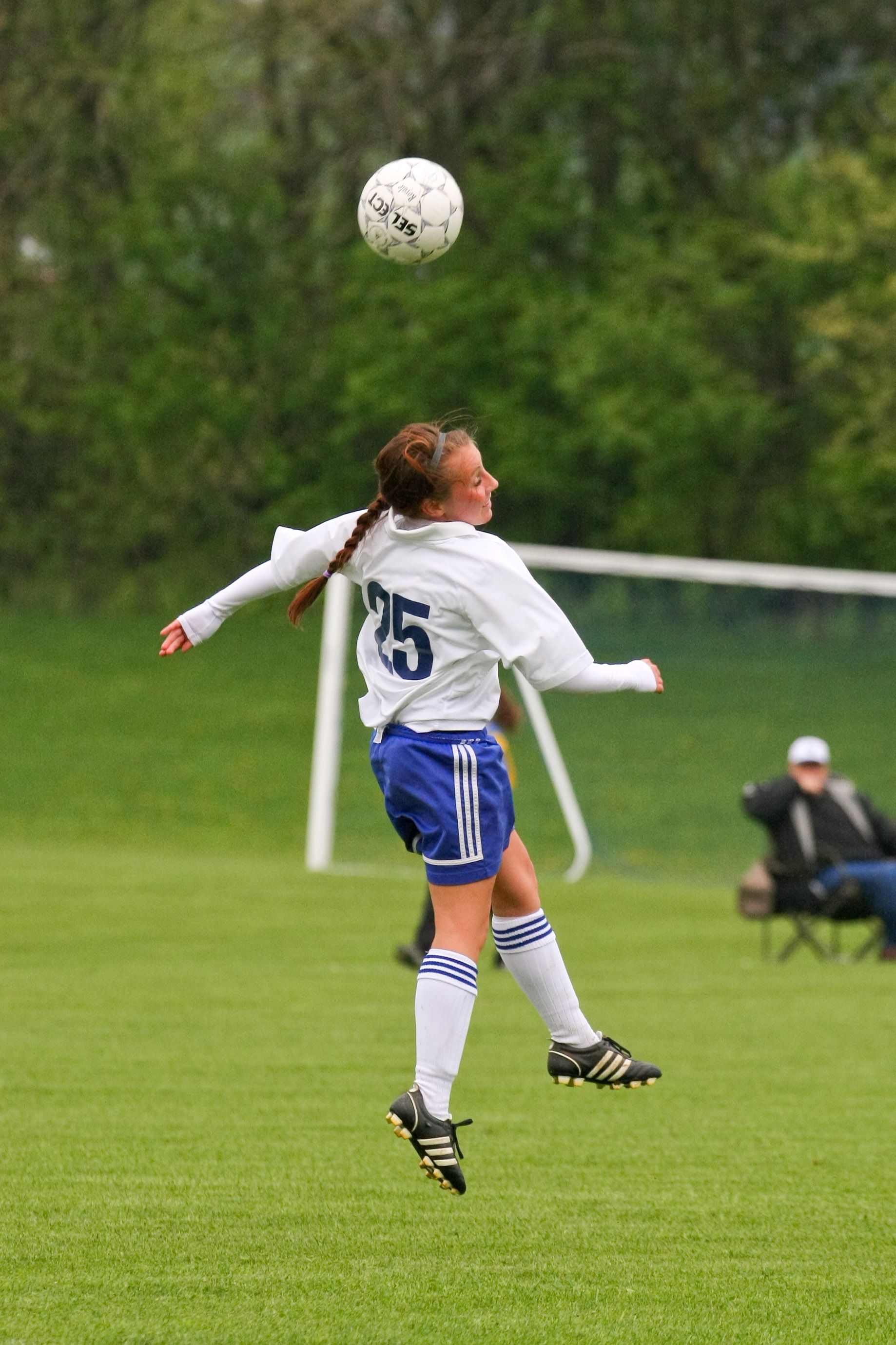Soccer player in white and blue jersey leaps for a header on a grassy field, ball overhead.