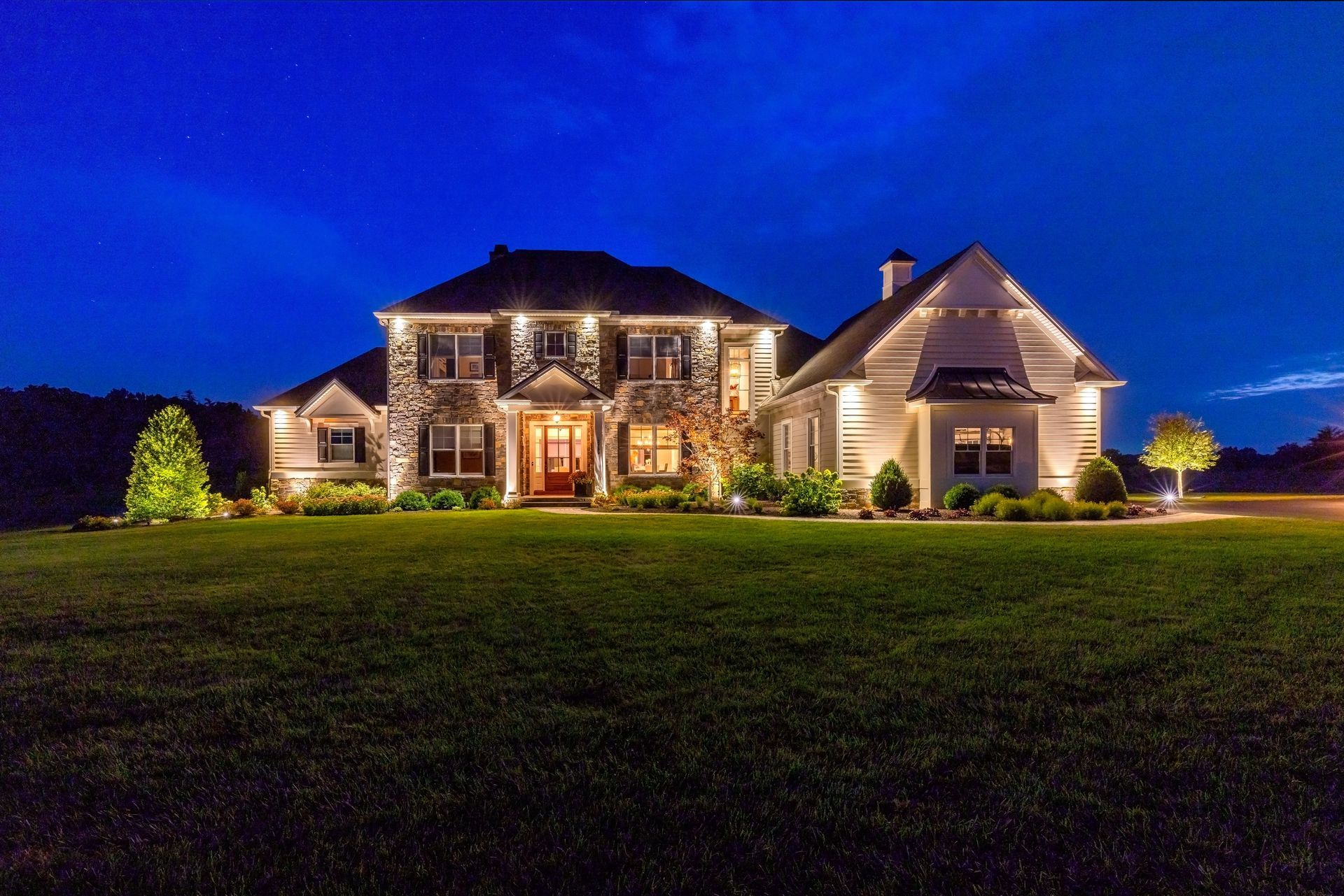 Large lit suburban house at dusk with a blue sky and a wide green lawn