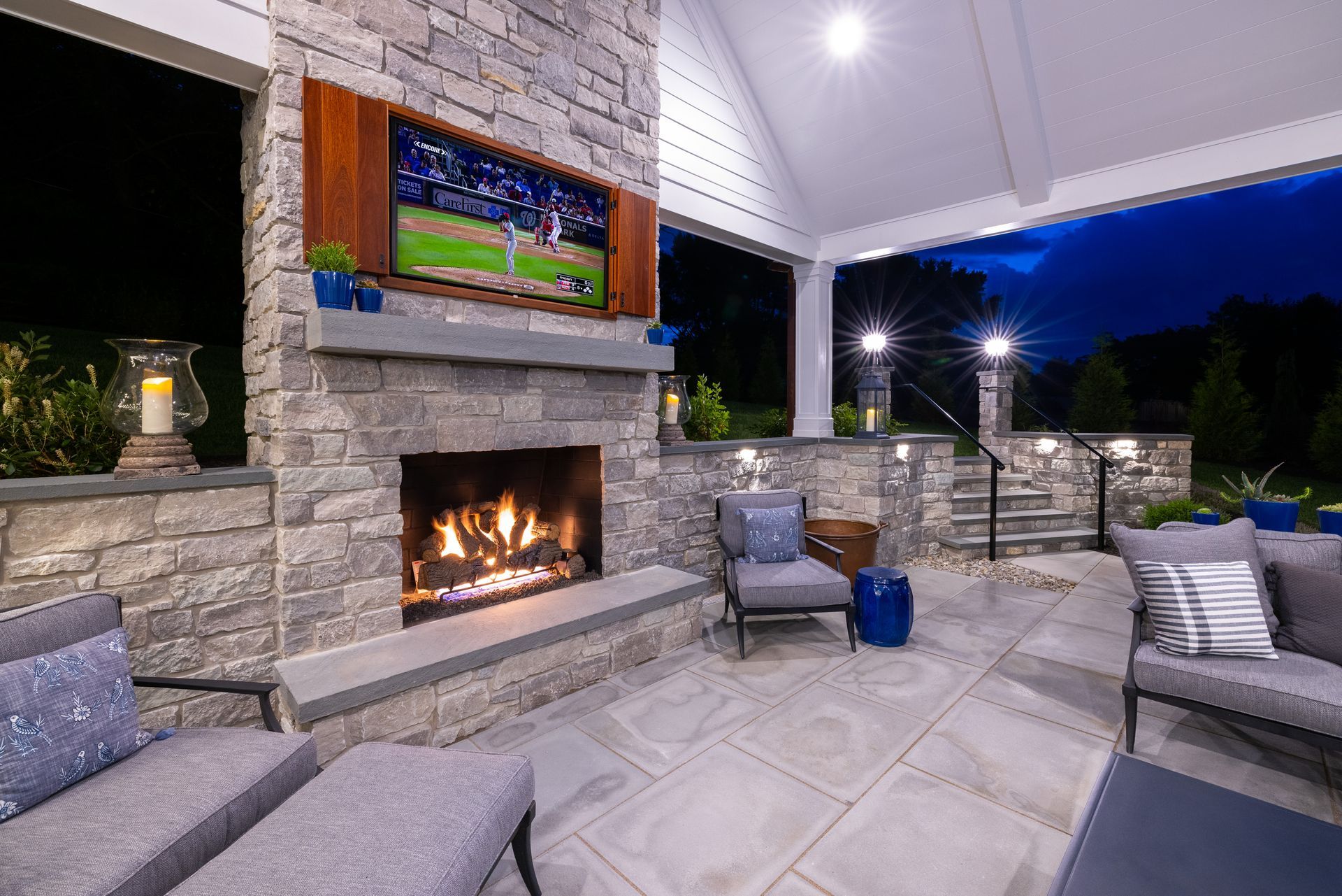 Covered patio with stone fireplace, wall TV, and cushioned seating at dusk
