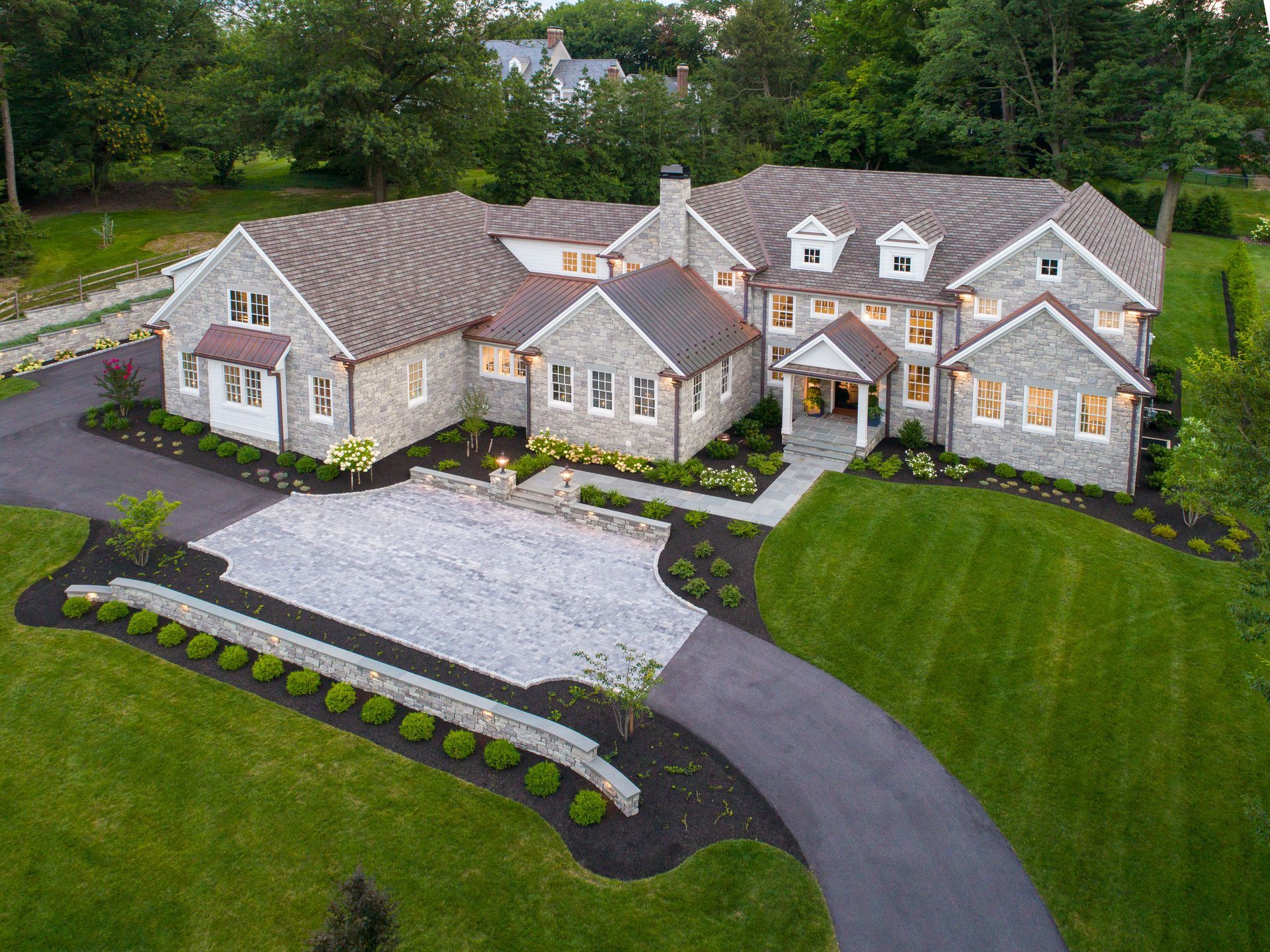Aerial view of a large gray stone house with a curved driveway, white gravel area, and manicured lawn.