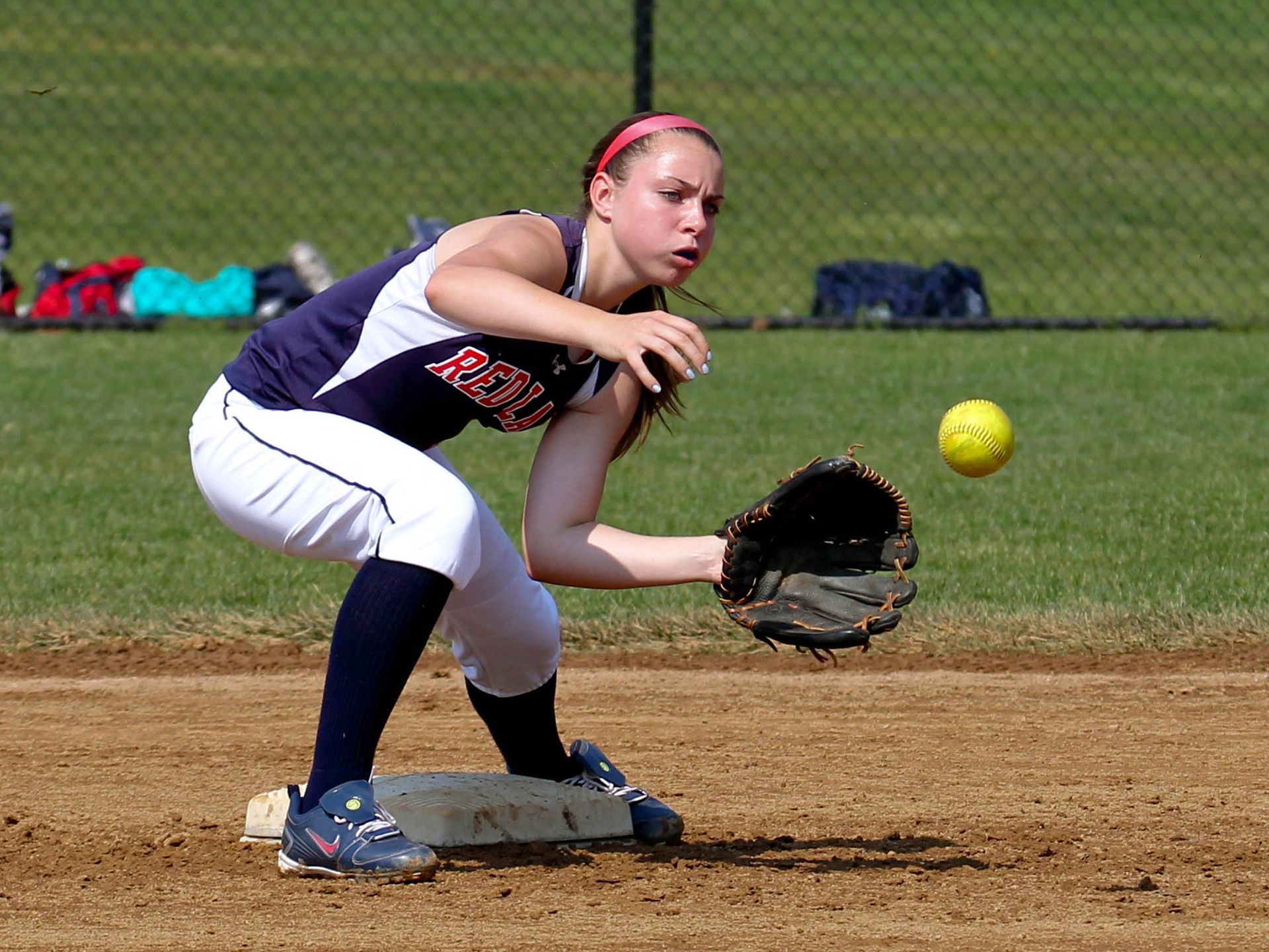Softball player crouches at base, glove ready for a yellow ball on the dirt infield.