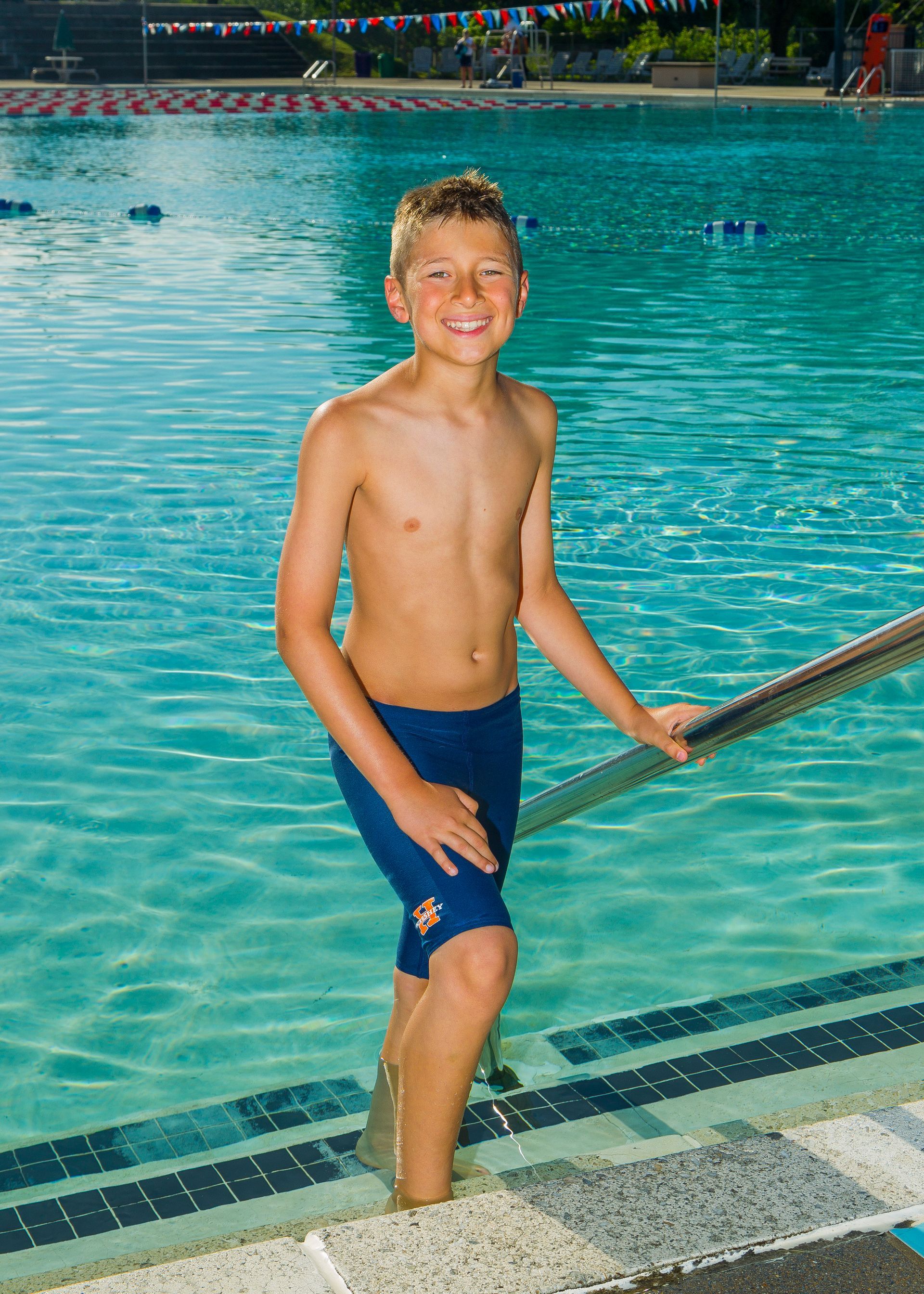 Smiling boy standing on pool steps in blue swim trunks by a swimming pool