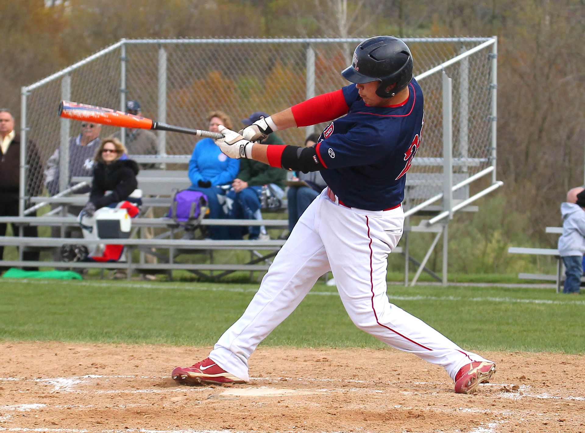 Baseball player in navy and red swings a bat on a dirt field with spectators in the background