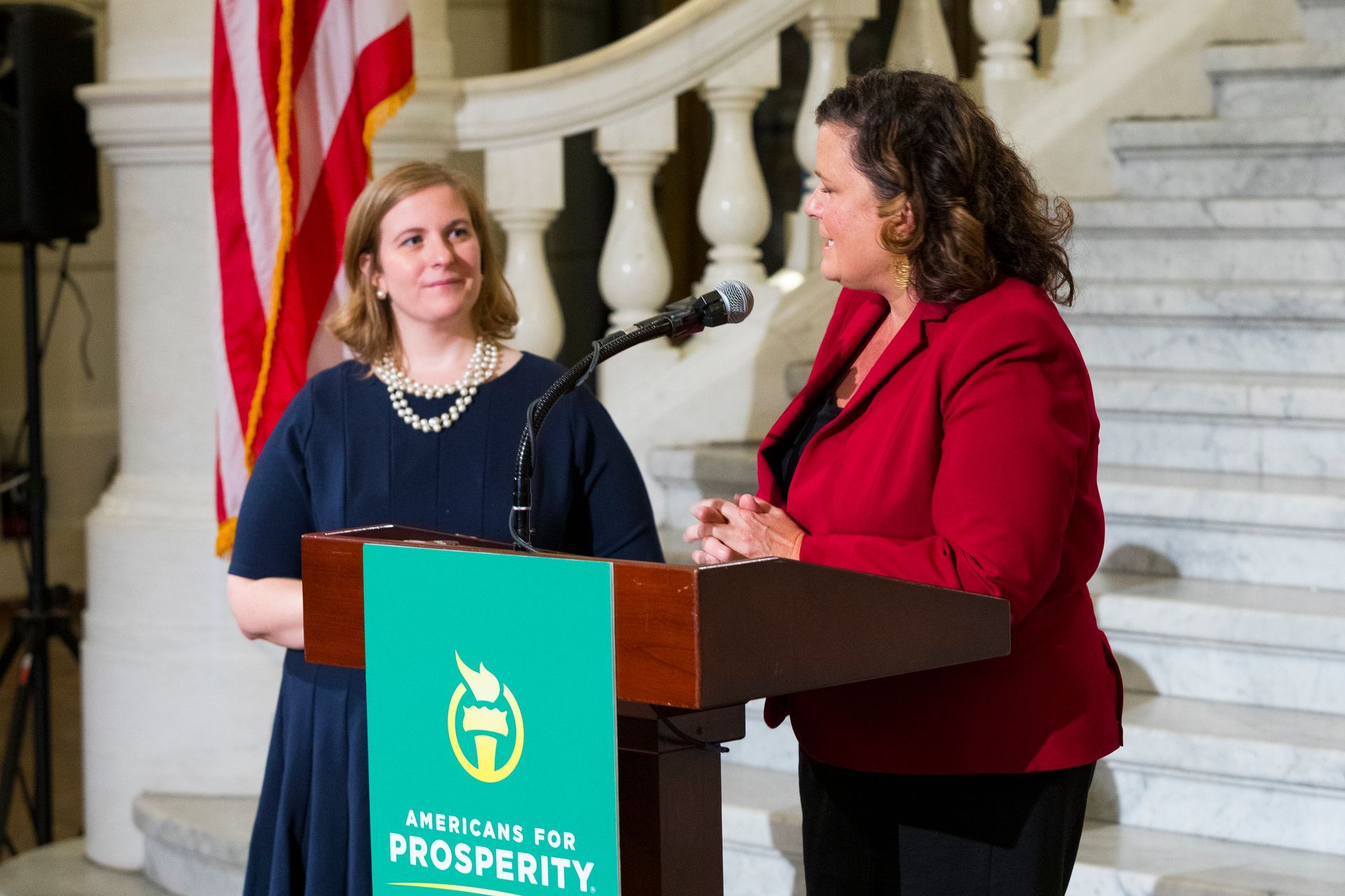 Two women speaking at a podium in a marble staircase hall with an American flag behind them