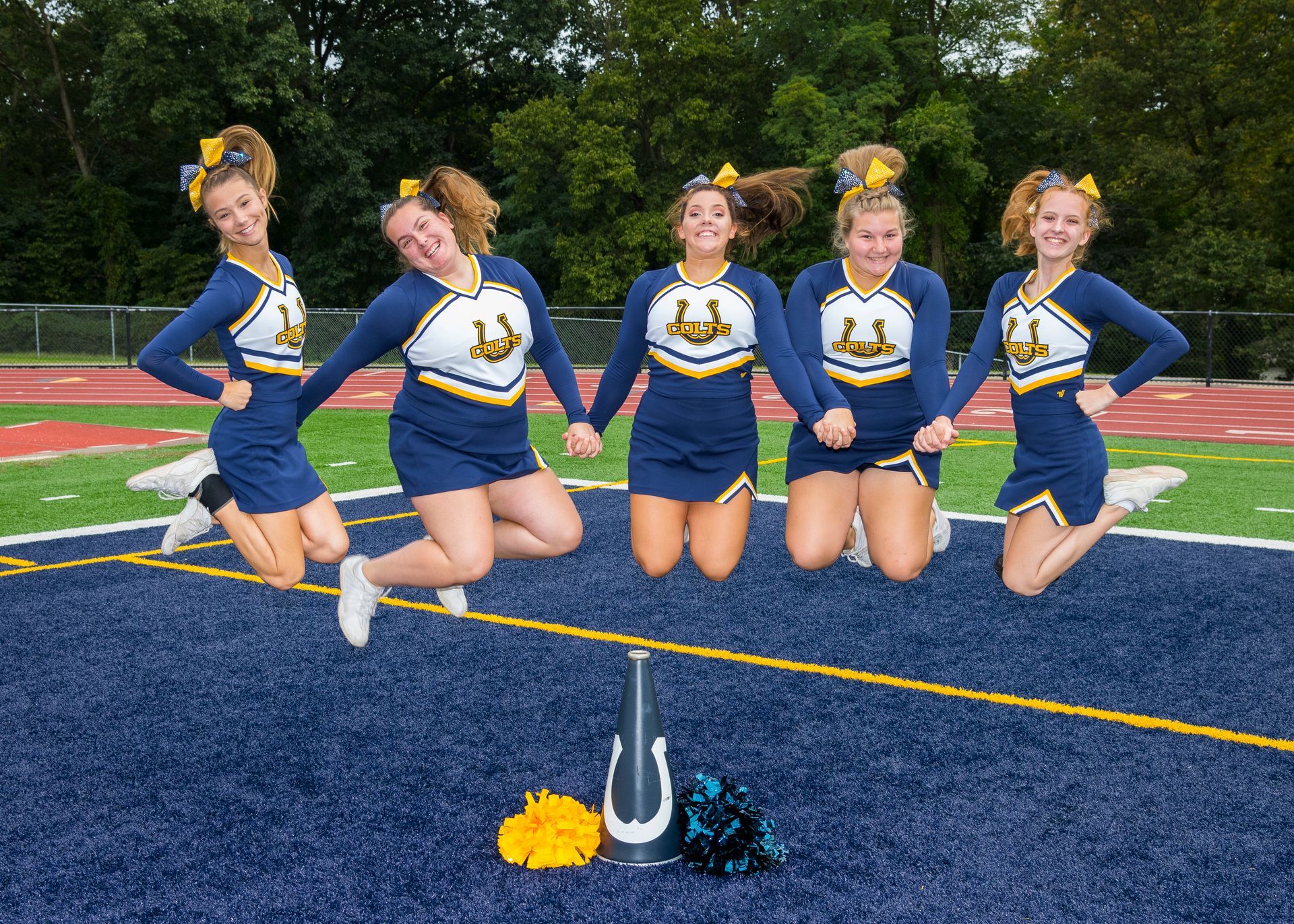 Five cheerleaders in blue uniforms pose on a football field with pom-poms in front.