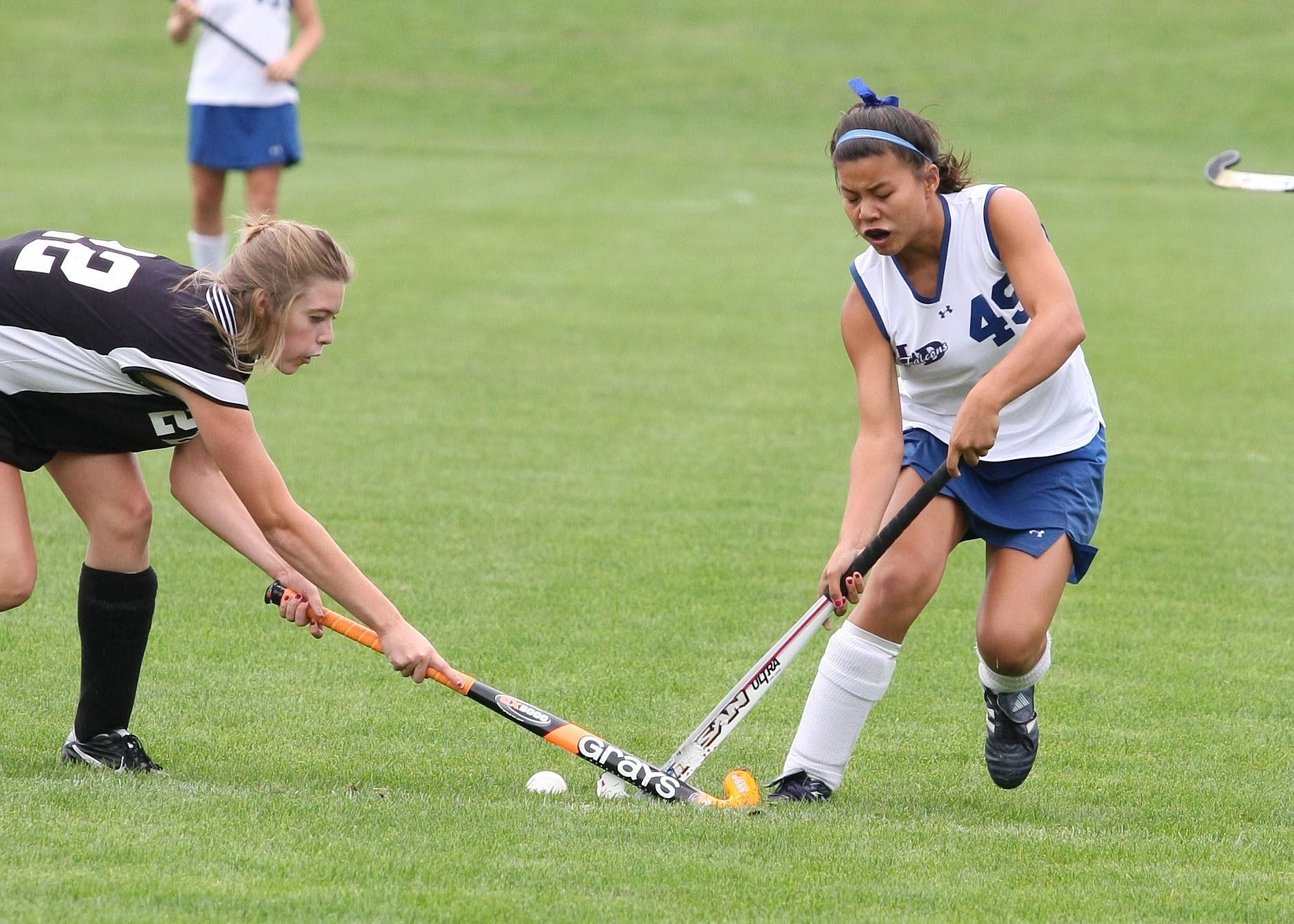 Two field hockey players in black and white uniforms contest the ball on a grassy field.