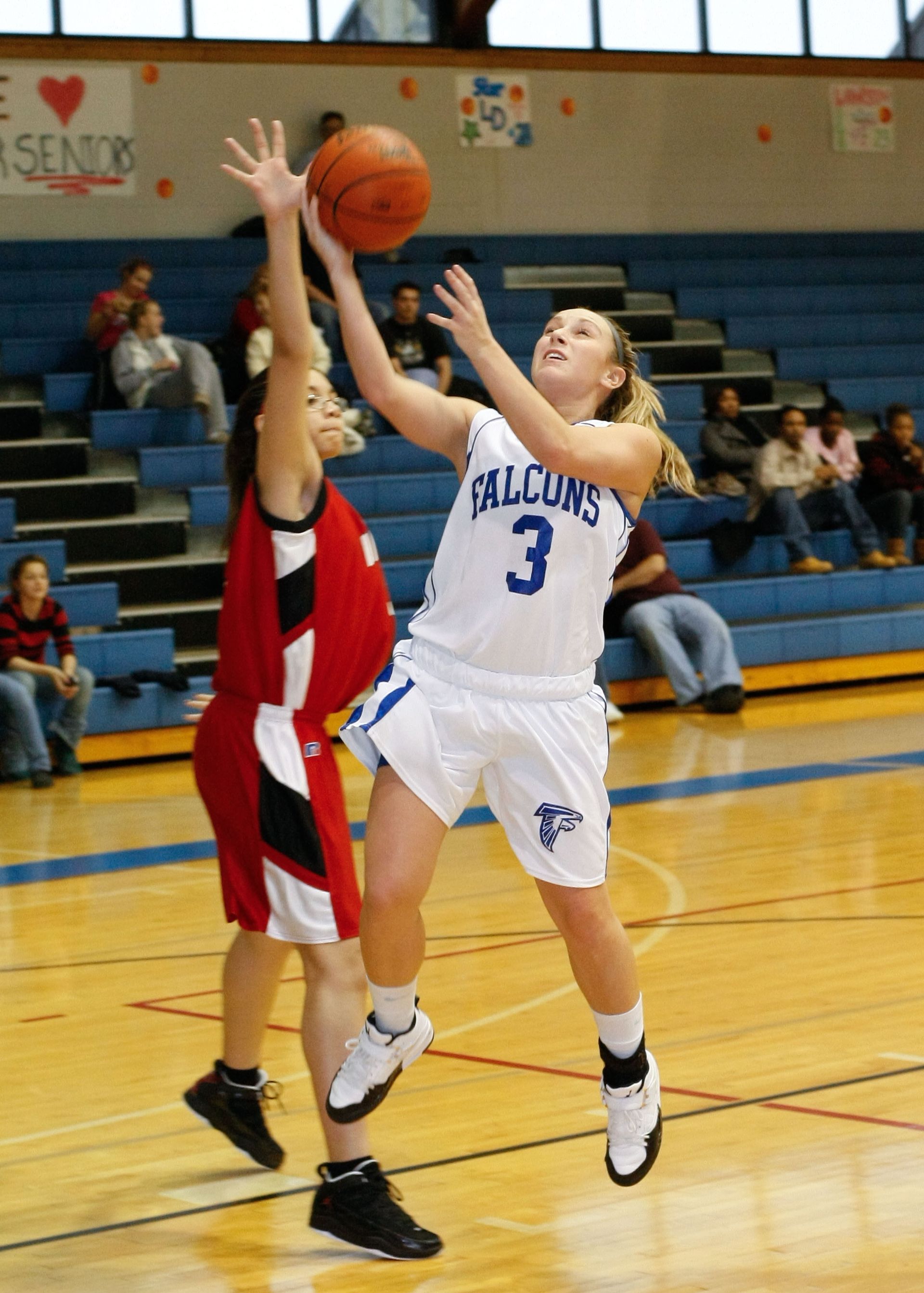 Two basketball players jump for a shot, one in white and one in red, on an indoor court.