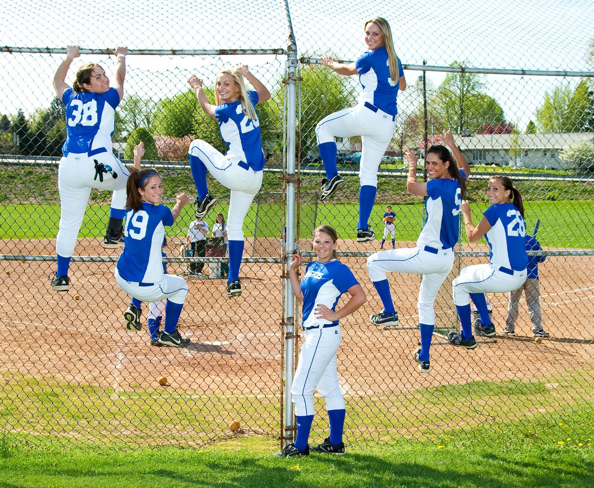 Softball team in blue jerseys posing in a circle on a dirt field behind a chain-link fence.