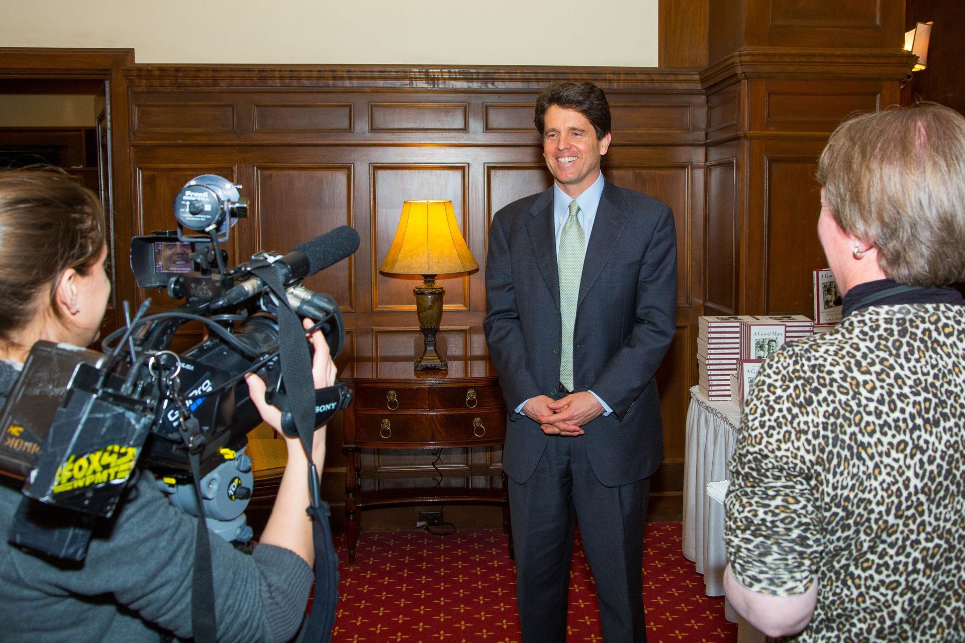 Man standing for a press interview in a wood-paneled room as cameras film him