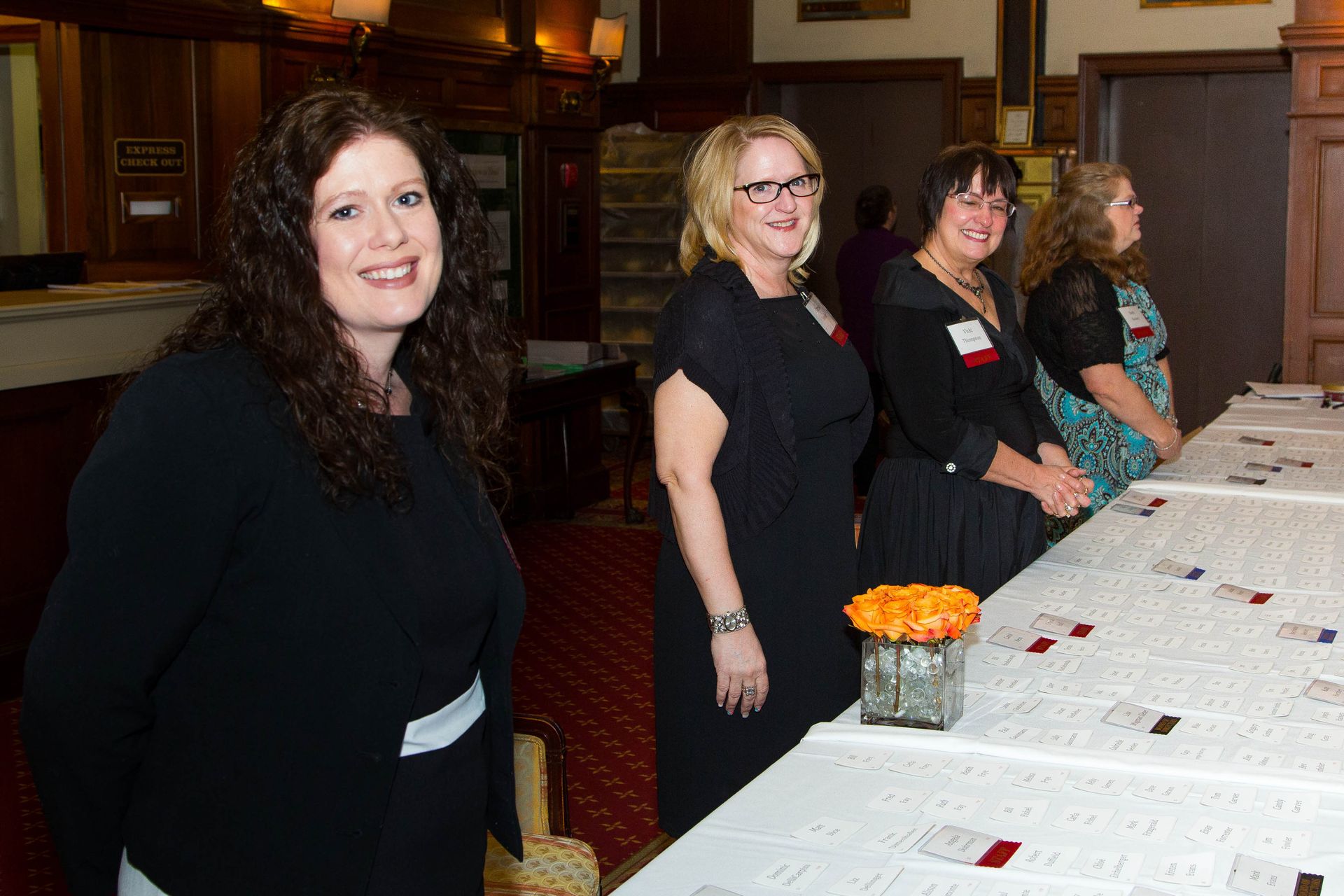 Four women standing behind a long table with name tags and flowers in a wood-paneled room