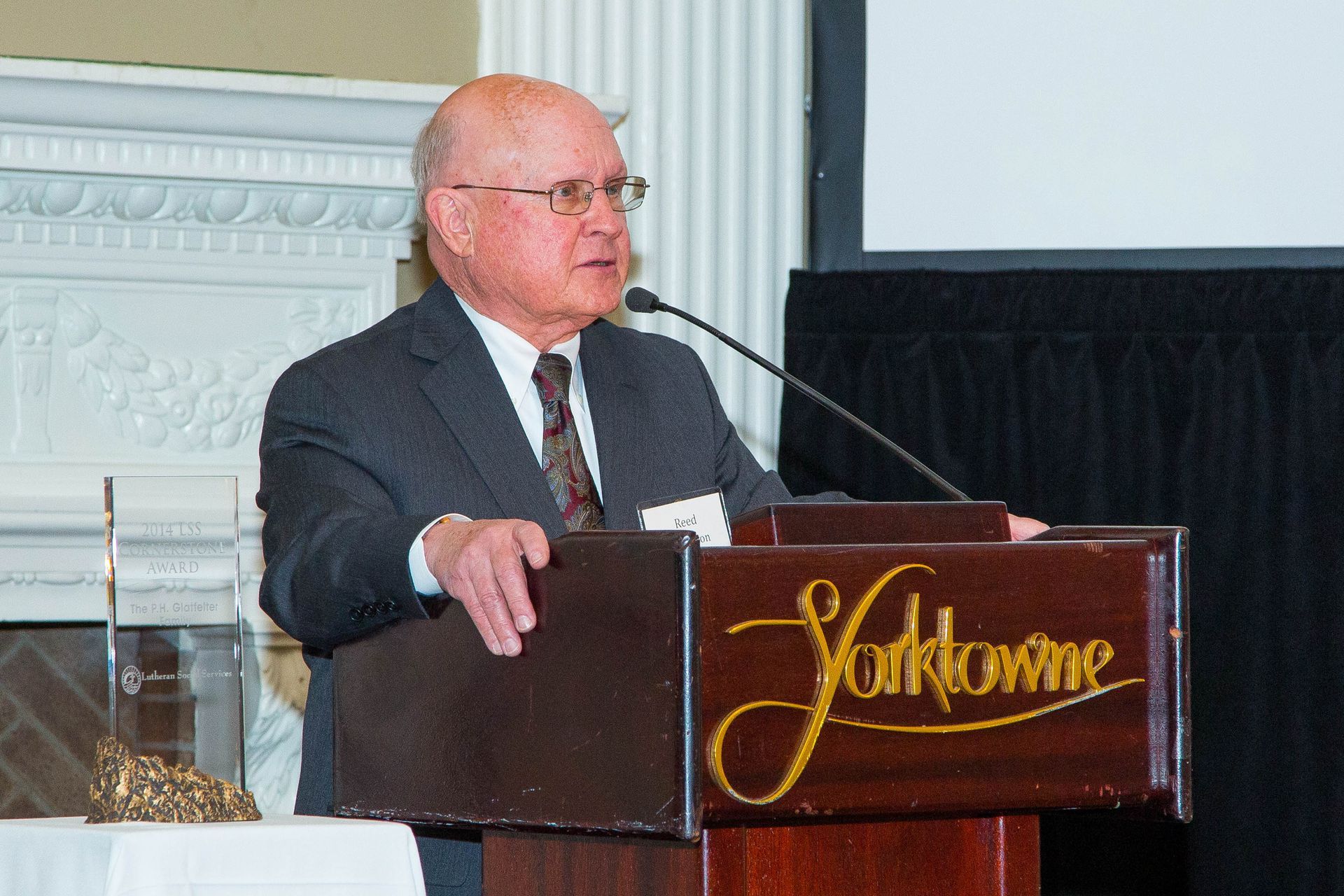Man speaking at a podium in a formal room, with a microphone and a gold sign on the front.